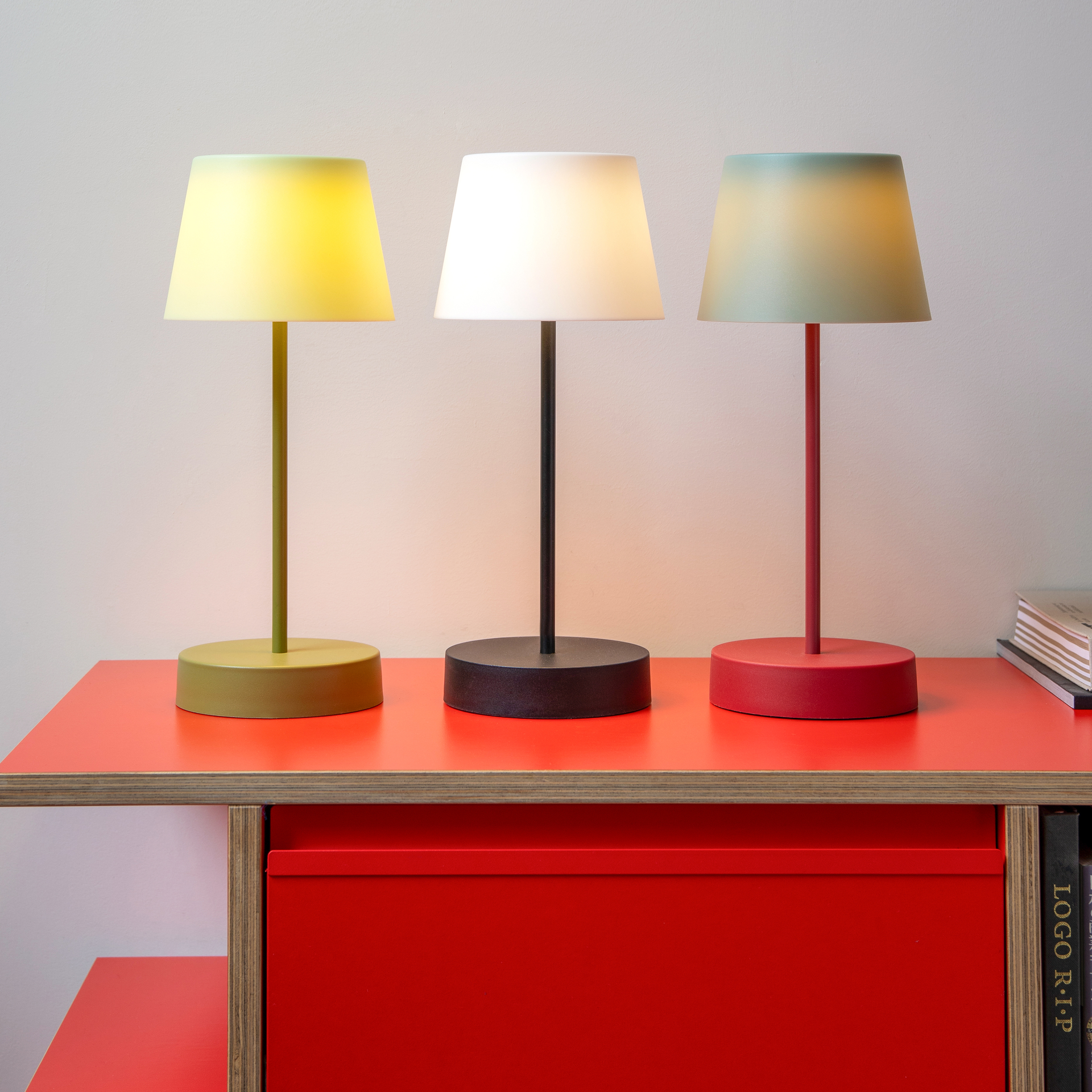 Drei verschiedenfarbige Tischleuchten der Serie Oscar auf einem roten Tisch. Die Lampen leuchten und haben einen runden Fuß.