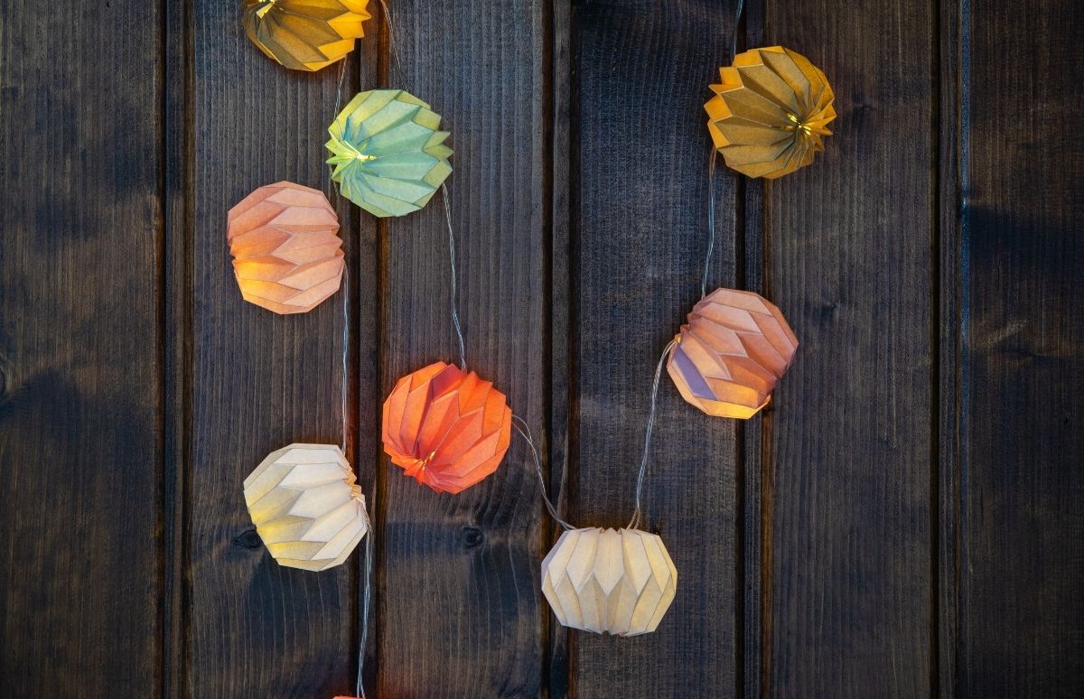 Eine vertikal hängende Lichterkette zeigt mehrere kleine, beleuchtete Papierlampions in herbstlichen Farben wie Gelb, Grün, Orange und Hellbraun. Sie leuchten sanft und werfen warmes Licht auf die dunkle, rustikale Holzwand.