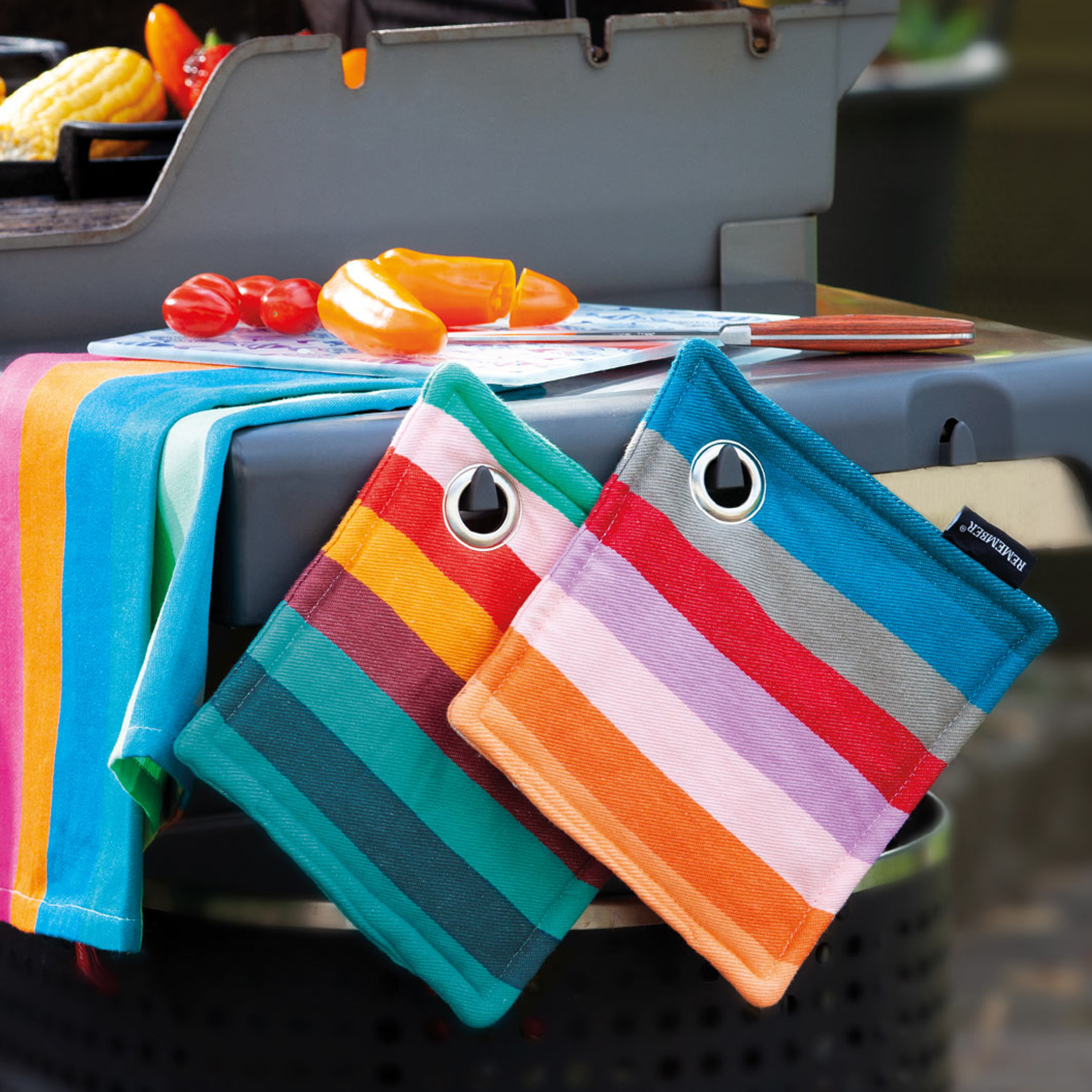 Zwei gestreifte Topflappen mit Aufhängeösen liegen auf einem Grill. Ein farblich passendes Geschirrtuch, Gemüse und ein Messer sind zu sehen.
