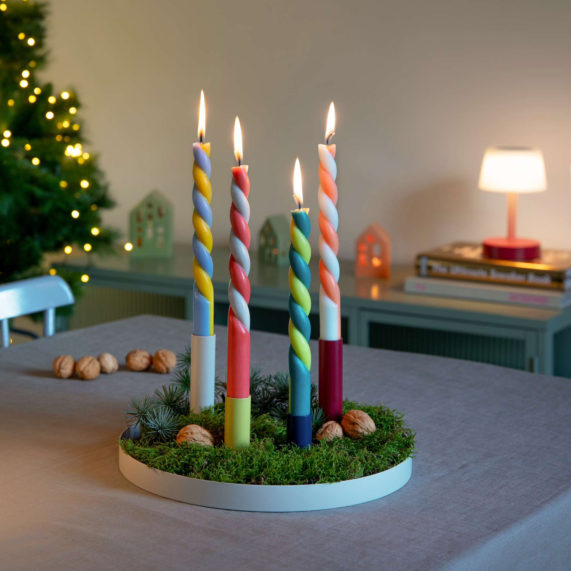 Quatre bougies torsadées multicolores sur un lit de mousse dans un plateau de bougies blanc. Les bougies sont allumées, avec un sapin de Noël illuminé en arrière-plan.