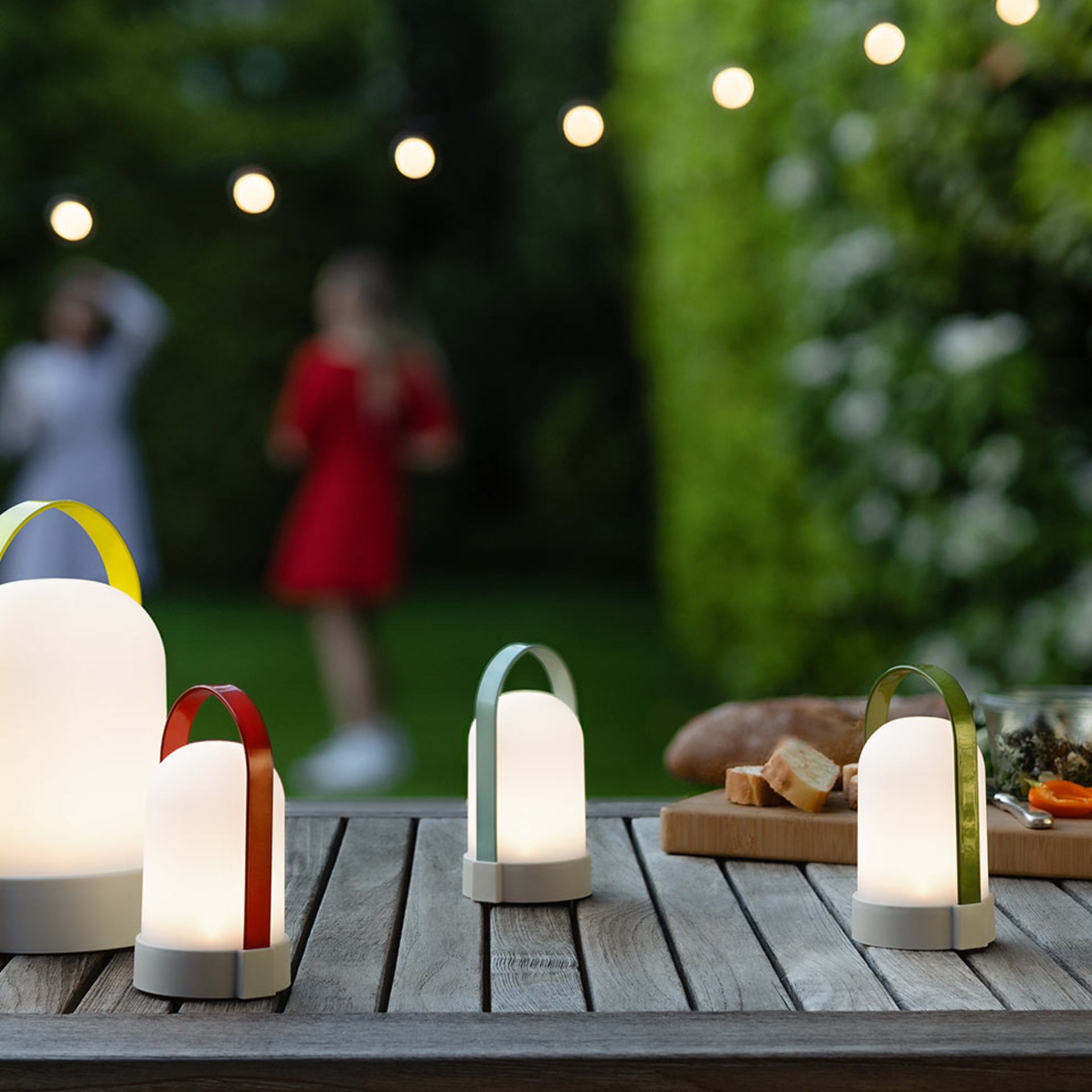 Drei Leuchten URI Piccolos mit farbigen Tragebügeln auf einem Tisch. Im Hintergrund sind Personen und Gartenbeleuchtung zu sehen.