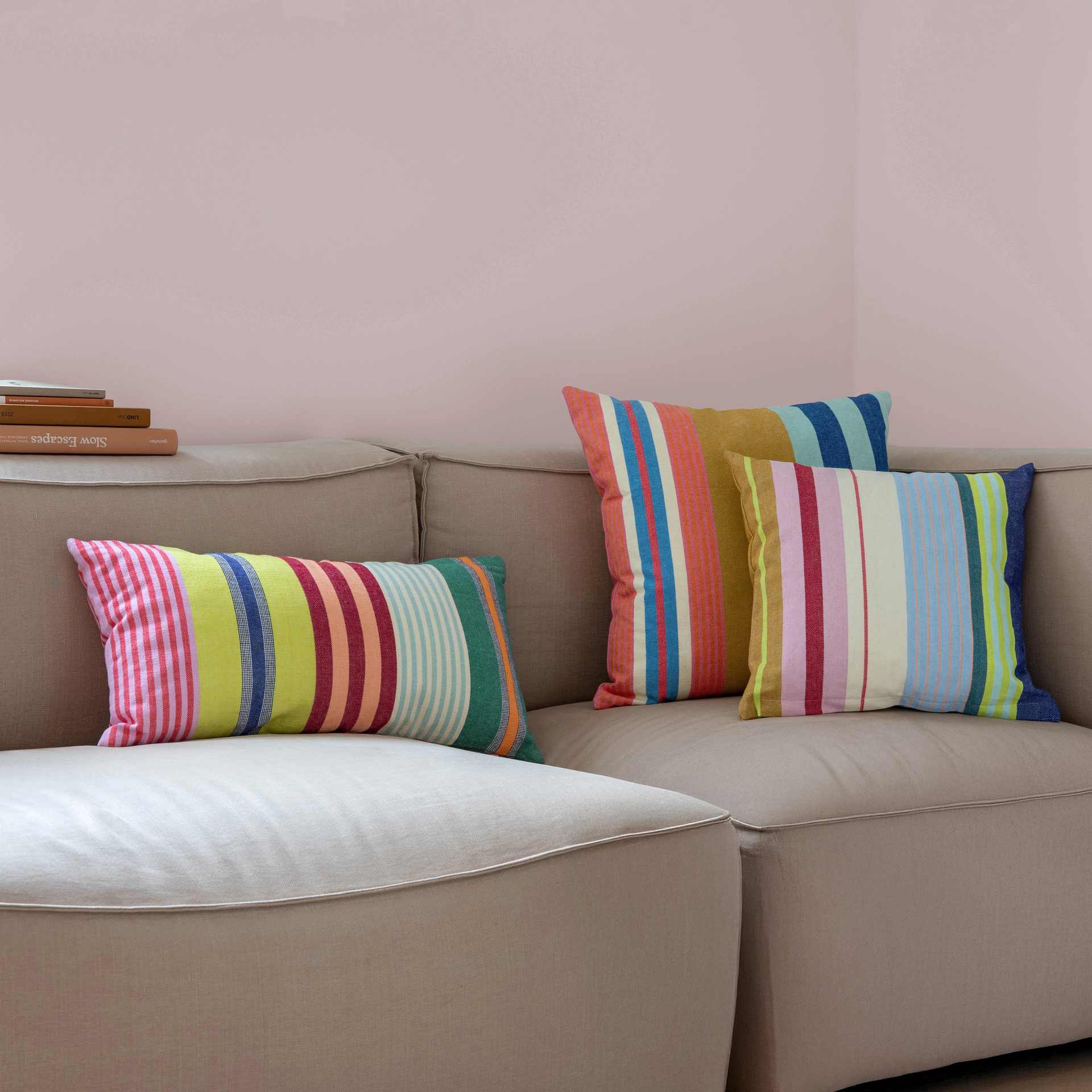 Nahaufnahme: Drei 'Belize' Baumwoll-Kissen mit bunten Streifen liegen auf einem beigen Sofa vor einer rosa Wand.