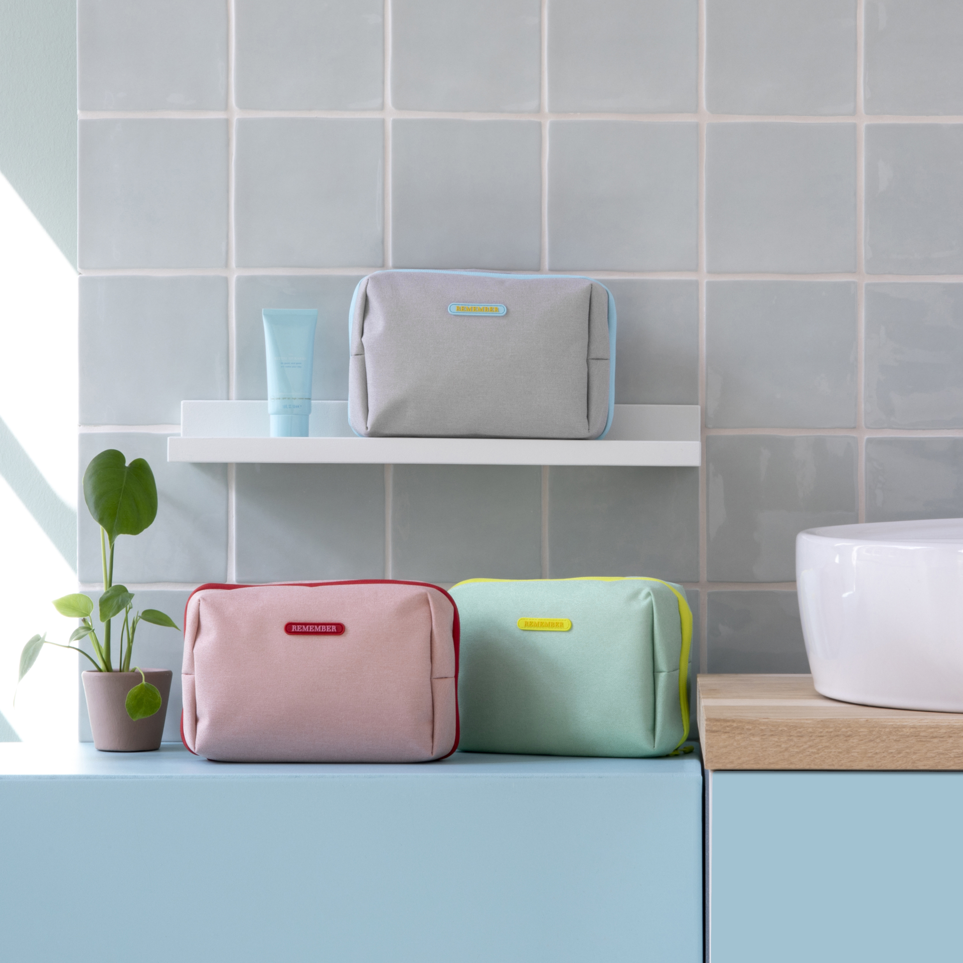 Drei Kulturtaschen in Grau, Rosa und Mint, jeweils mit farblich passendem Reißverschluss und Logo, stehen im Bad. Dazu ein Töpfchen mit einer Pflanze und eine Tube.