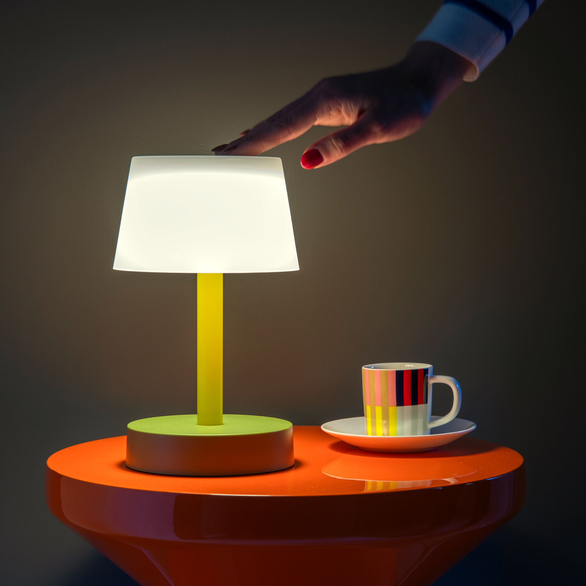 Eine Hand berührt den Lampenschirm der Tischleuchte Fritz 'flora'. Das Produkt, mit gelbem Stab und grünem Fuss, steht auf einem orangen Tisch. Daneben steht eine Tasse.