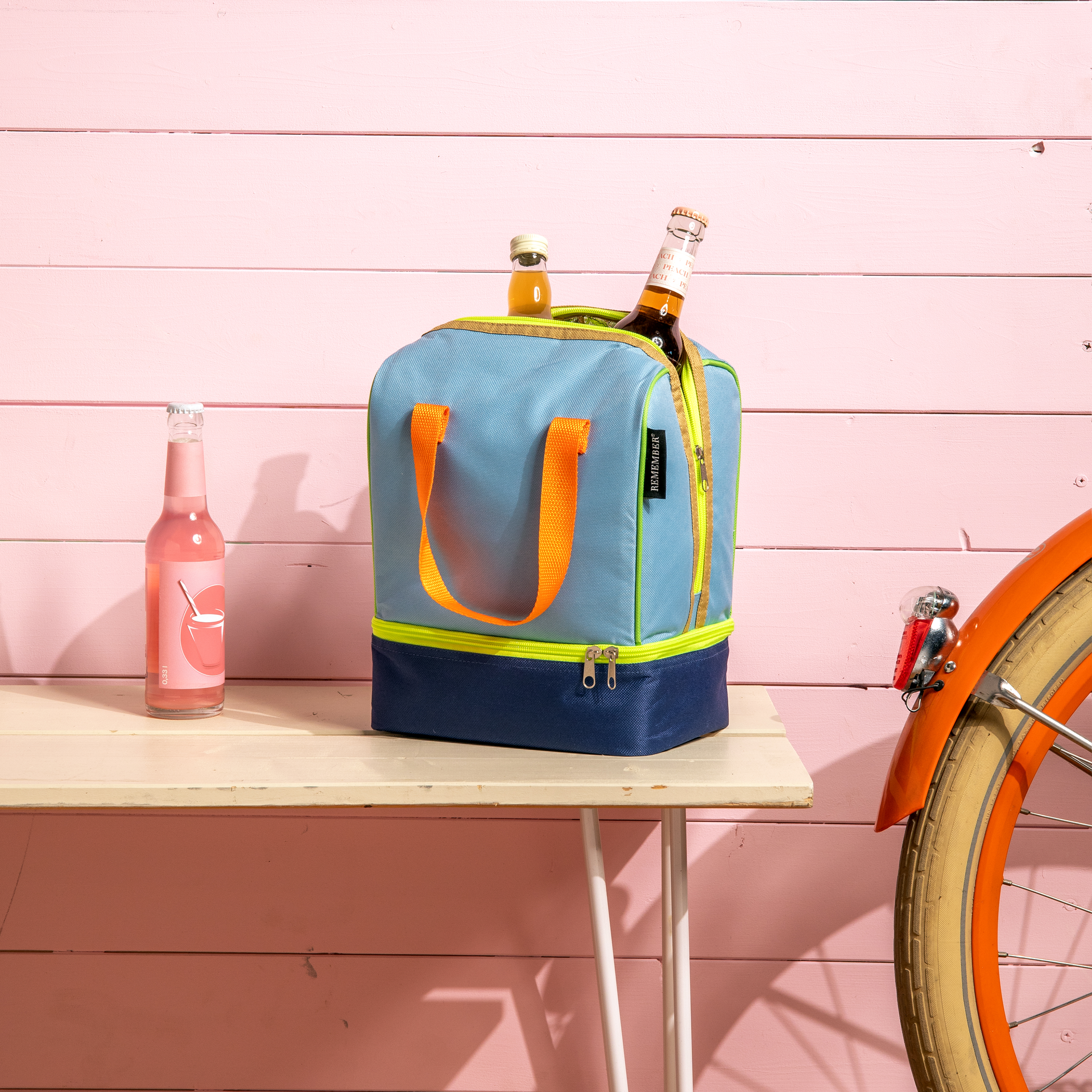 Hellblaue Lunch-Kühltasche 'Nick' mit orangen Trägern, gefüllt mit Getränkeflaschen, steht auf einem Tisch. Im Hintergrund eine rosa Wand und ein Fahrrad.