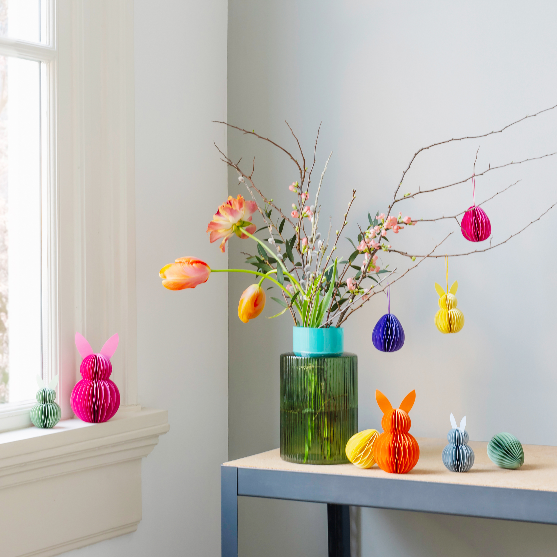 Dekorative Ostereier und Hasen Baumschmuck aus Papier stehen neben einer Vase mit Zweigen und Blumen auf einem Tisch. Einige der Dekorationen hängen an den Zweigen.
