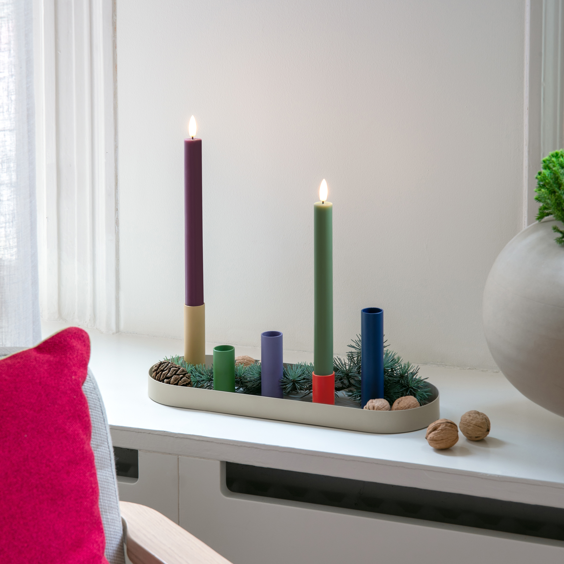 Zwei LED-Stabkerzen, eine olivgrün und eine auberginefarben, stehen neben einer weißen Fernbedienung mit bunten Knöpfen.