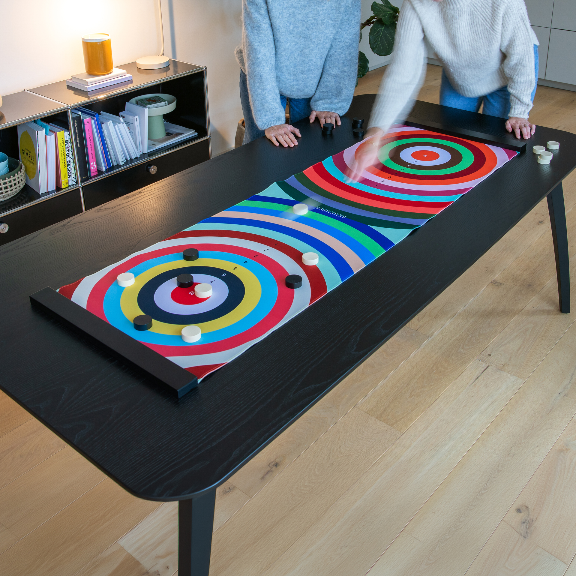 Zwei Personen spielen ein Tischspiel mit bunten Zielkreisen auf einer schwarzen Tischplatte. Schwarze und weiße Spielsteine liegen verstreut auf dem Spielfeld.