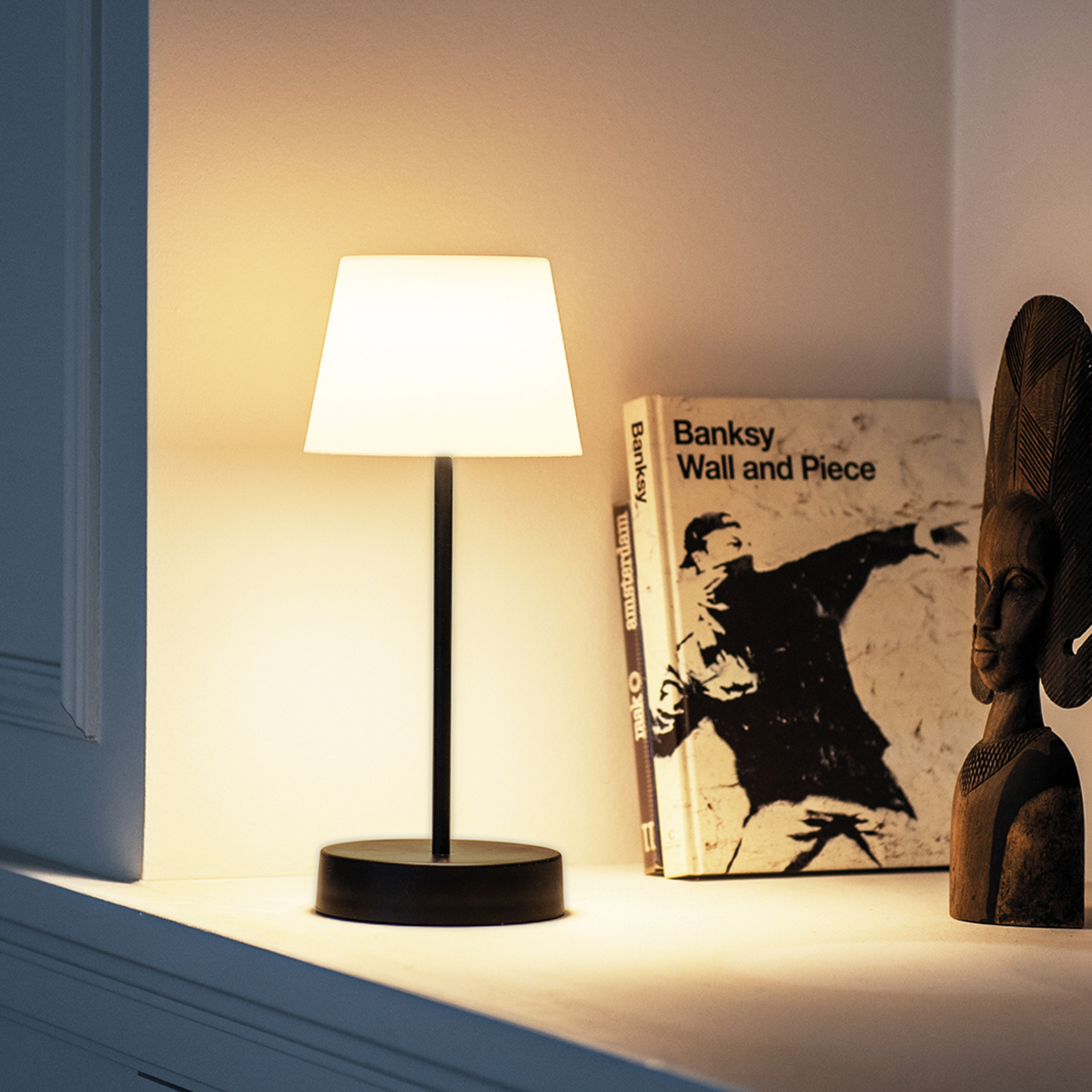 Die Tischleuchte Oscar 'pure' steht auf einem Sims. Die Lampe hat einen schwarzen Fuß und Stab, der Lampenschirm ist hell. Ein Buch und eine Holzfigur stehen daneben.