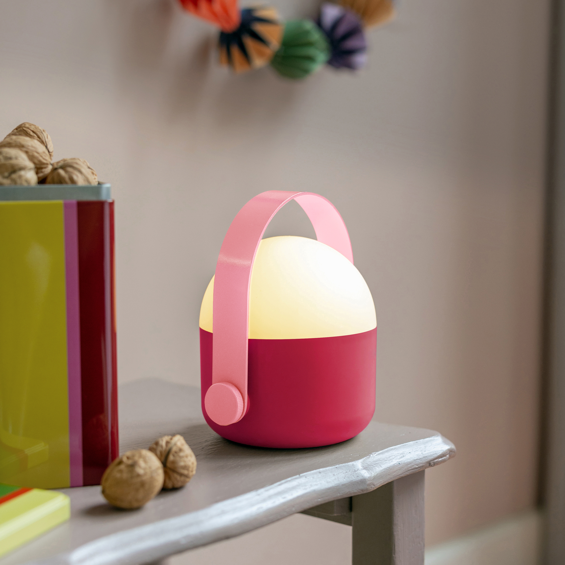Die Leuchte Ole in Berry: Rote, tragbare LED-Lampe mit rosafarbenem Henkel. Der obere Teil ist weiß und leuchtet. Seitlich ein runder Knopf zum Dimmen.