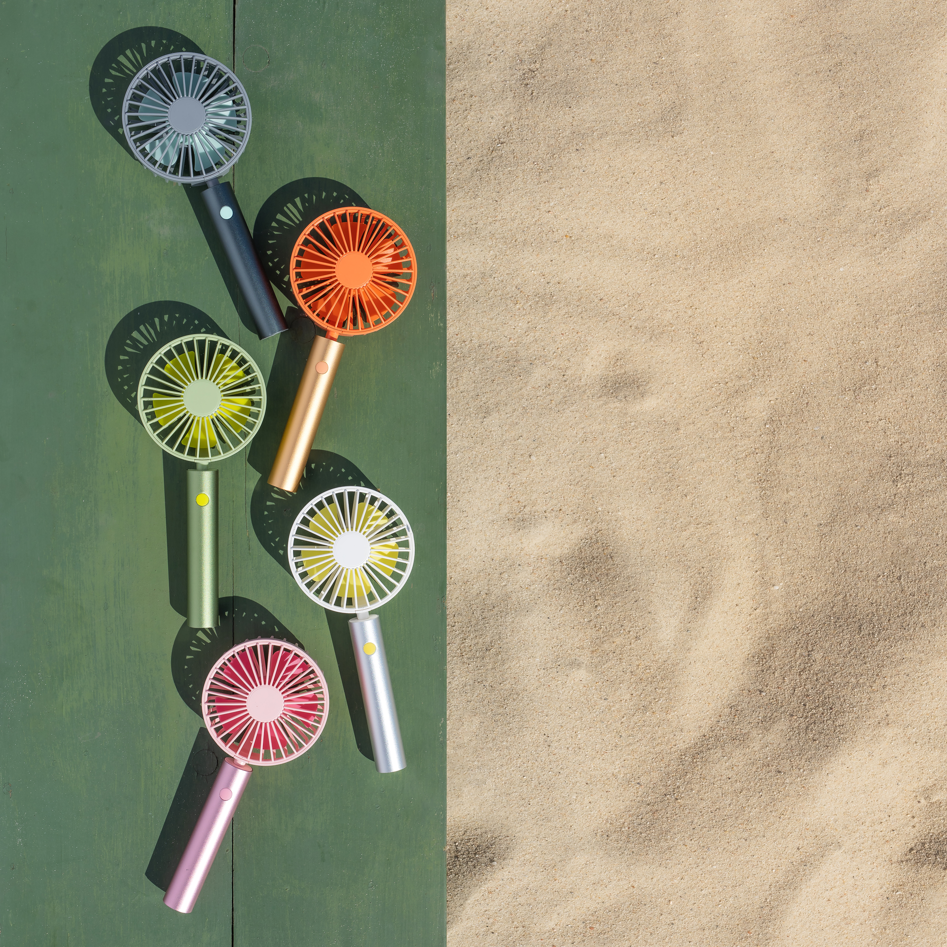 Mehrfarbige Flow Ventilatoren auf Grün und Sand, Draufsicht. Die Ventilatoren sind in Grau, Orange, Gelb, Weiß und Pink.