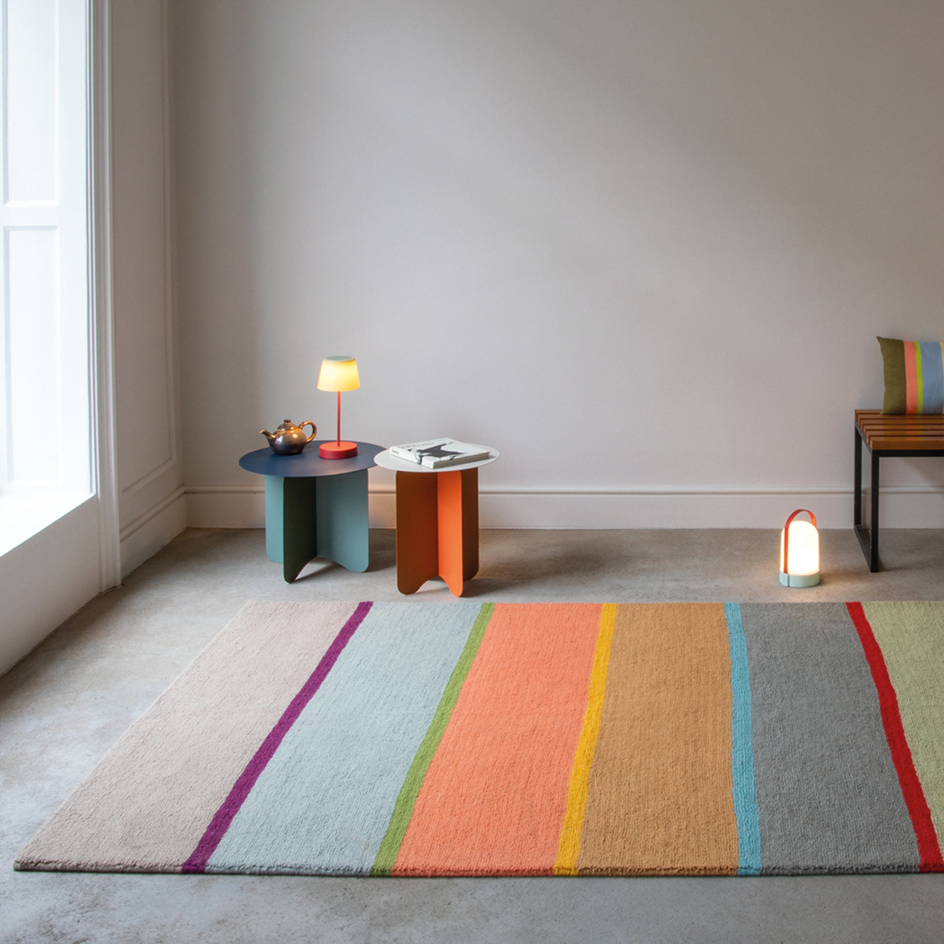Un tappeto "Cambridge" a strisce colorate si trova in una stanza luminosa. Tavoli decorativi, lampade e sedute con cuscini completano l'arredamento.