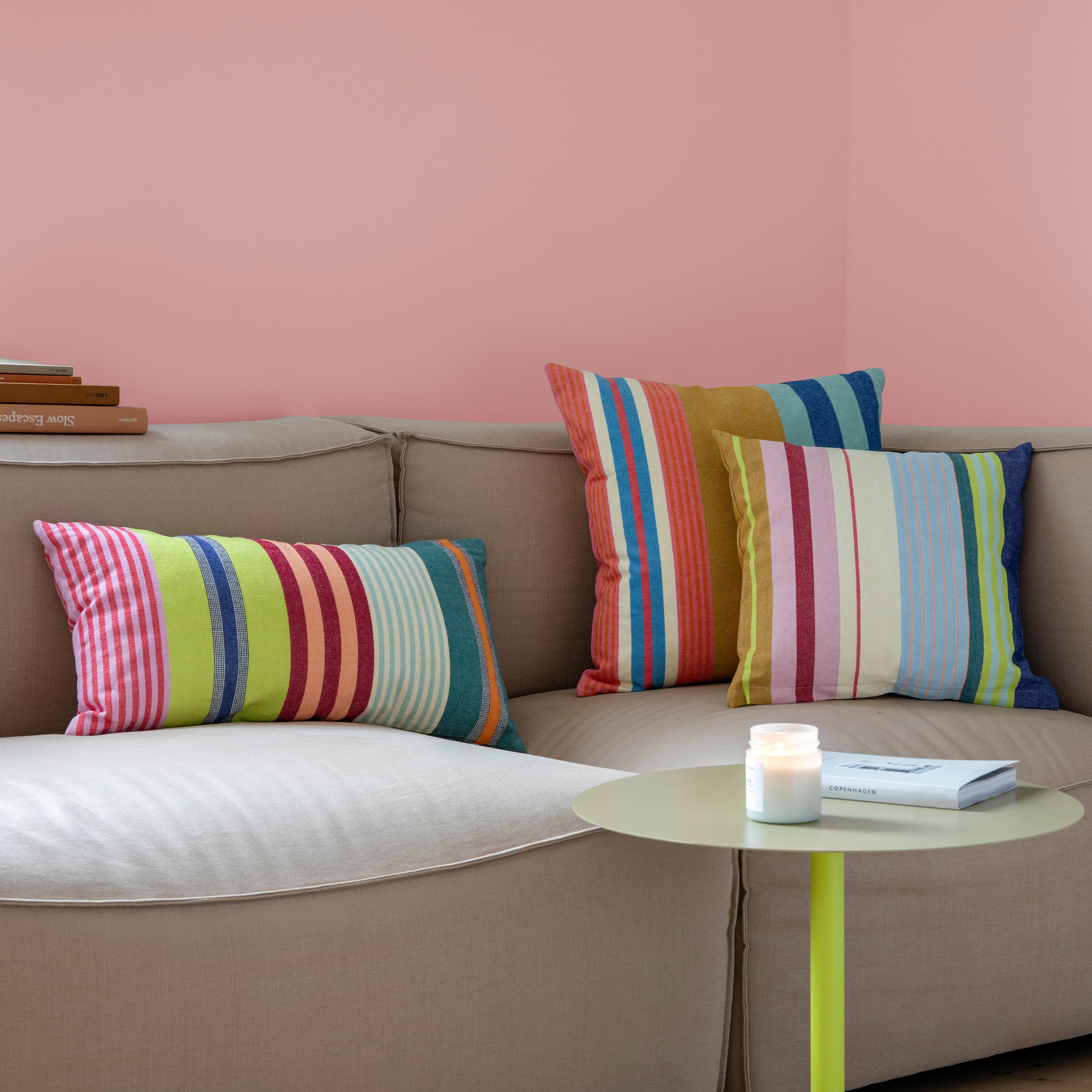 Beige Sofa mit drei gestreiften Baumwoll-Kissen 'Barbados' in bunten Farben. Ein kleiner, runder Tisch mit Buch und Kerze steht davor, die Wand ist rosa.