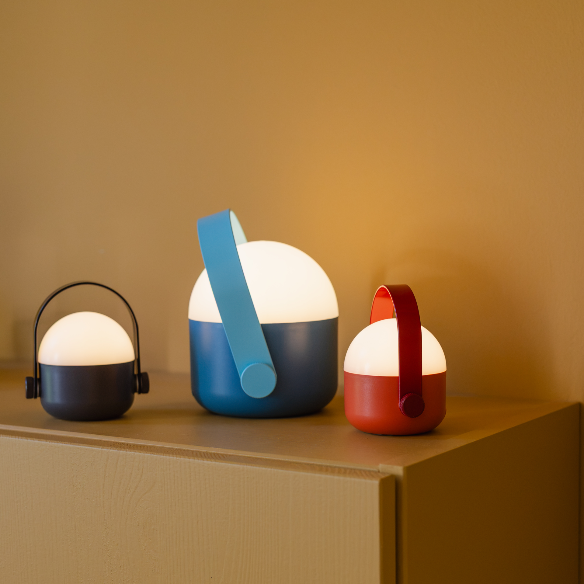 Drei leuchtende, akkubetriebene OLE mini LED-Leuchten stehen auf einer Holzkommode vor brauner Wand. Links klein, dunkelgrau mit schwarzem Bügel; mittig größer, dunkelblau mit hellblauem Bügel; rechts klein, rot. Warmweißes Licht, mit Griff.