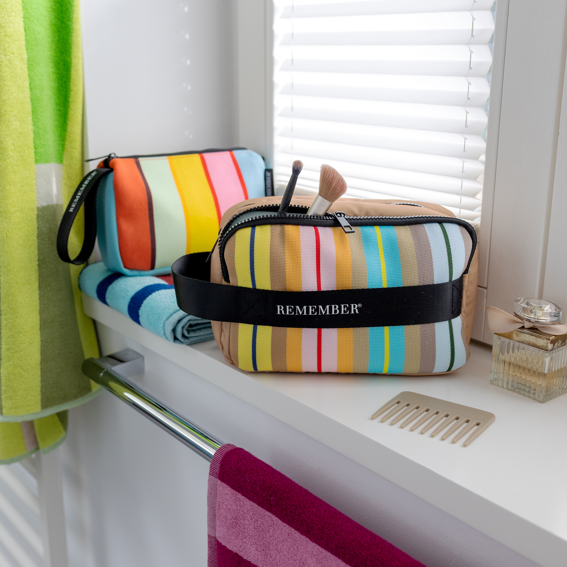 Deux trousses de toilette colorées et rayées sont posées sur une étagère blanche. La trousse Ventara au premier plan est ouverte et contient des brosses. Derrière, une autre trousse repose sur une serviette, près d’un peigne.