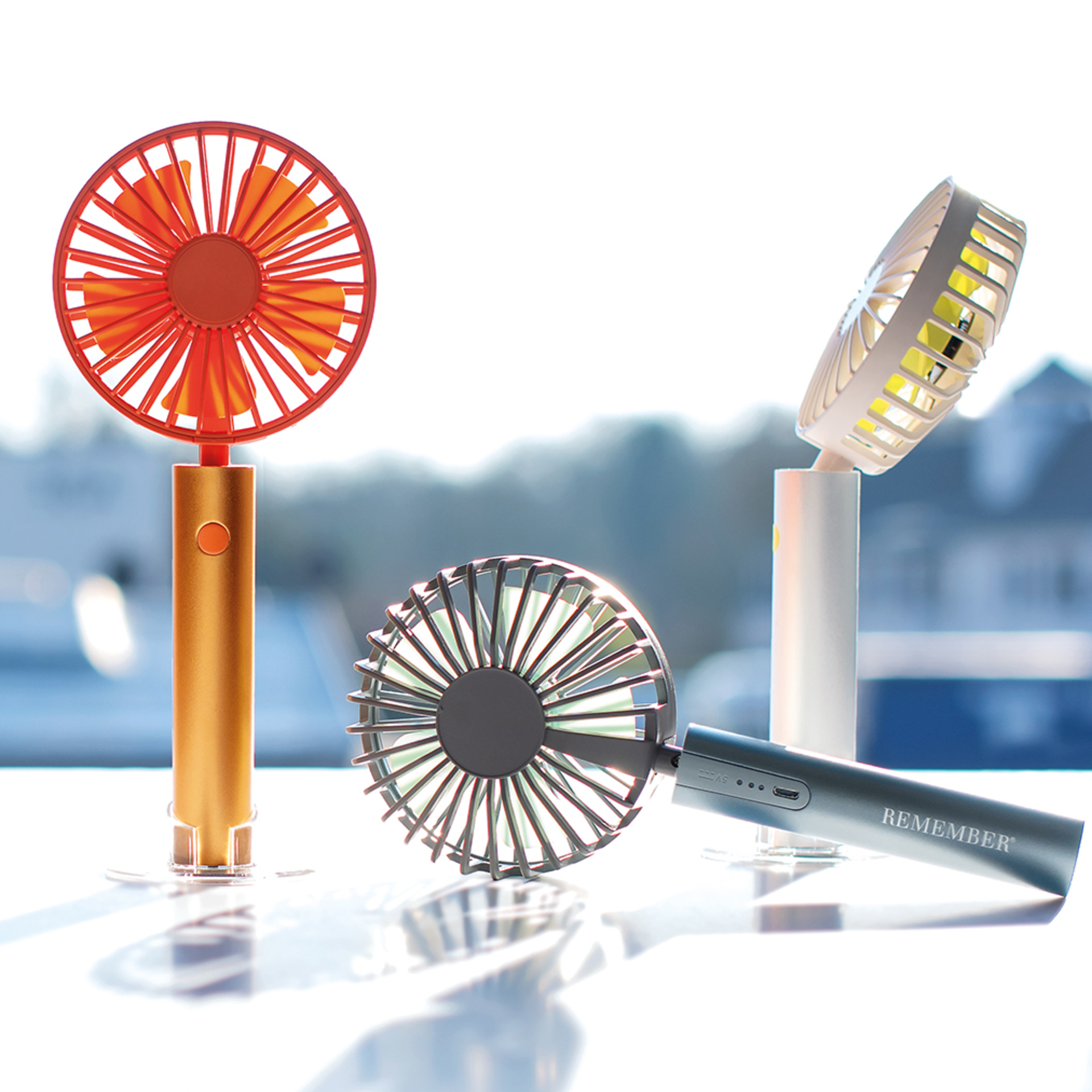 Quattro ventilatori a batteria nei colori arancione, grafite, grigio e bianco con base trasparente su sfondo chiaro. Un ventilatore è sul lato.