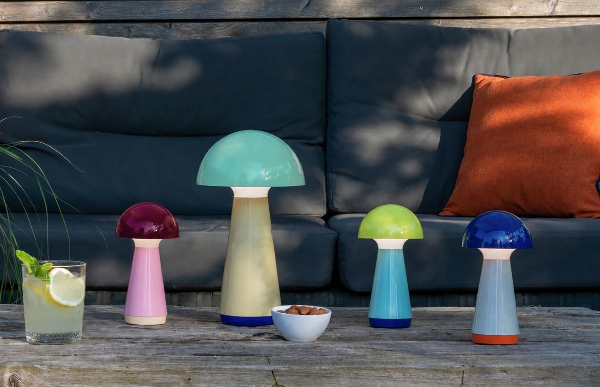 Vier leuchtende pilzförmige LED-Tischlampen in Rot, Türkis, Grün und Blau auf Holztisch. Links ein Glas Limonade mit Minze und Zitrone, rechts eine Schale Mandeln. Im Hintergrund ein dunkles Sofa mit orangem Kissen.