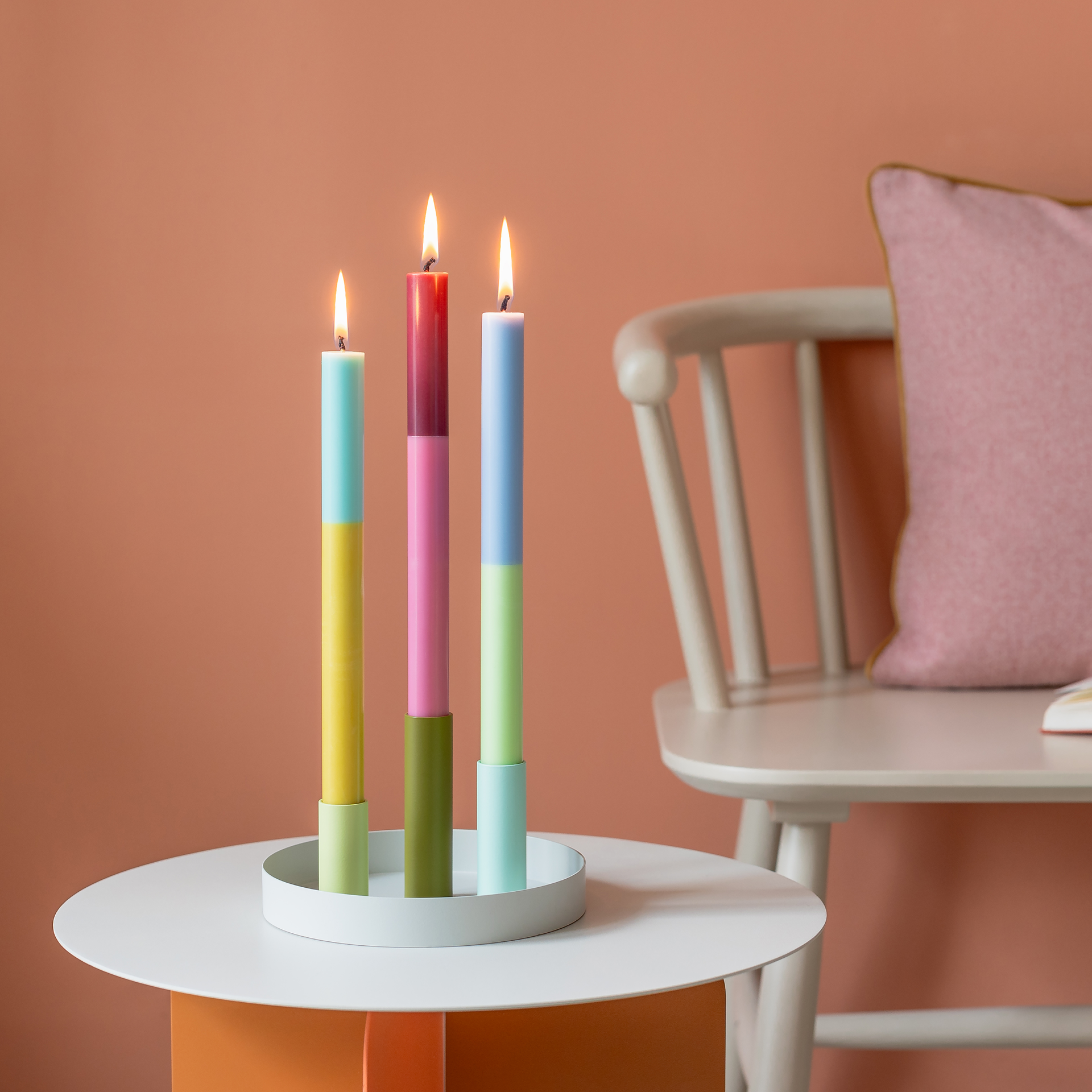 Trois bougies bâton allumées et colorées au design color-block sont disposées sur une table. Les bougies sont disposées dans un plateau à bougies blanc. En arrière-plan, une chaise et un mur.