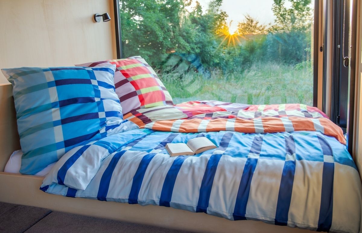 Un letto accogliente, con coperte a quadretti blu e bianchi e a strisce arancioni e rosse, si trova davanti a una grande finestra che si affaccia su un paesaggio verdeggiante all’alba o al tramonto. C’è un libro aperto a disposizione.