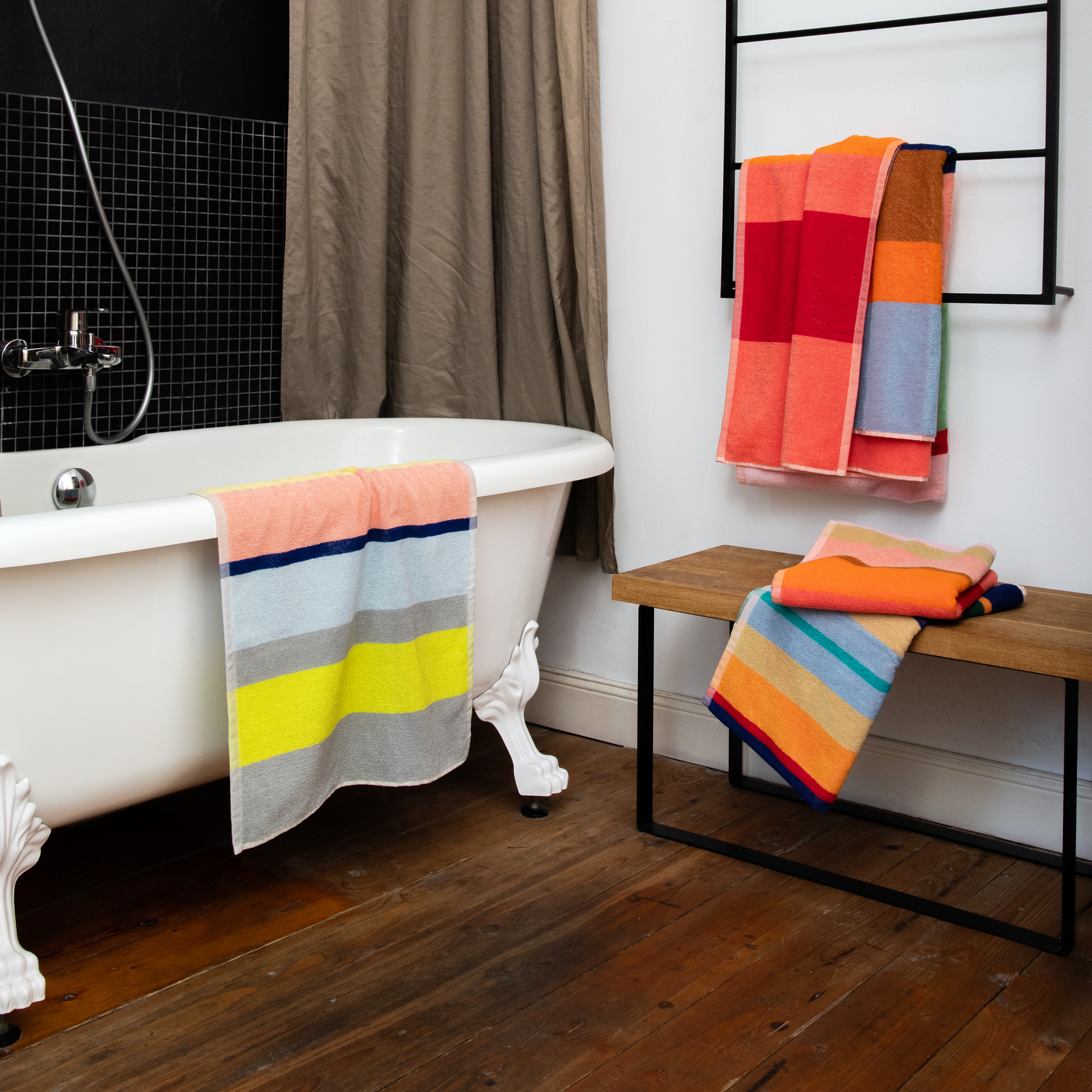 Ein Badezimmer mit Badewanne, Handtüchern und Bank. Ein gestreiftes Handtuch hängt über dem Rand der Badewanne. Weitere Handtücher sind auf einer Bank und einem Handtuchhalter platziert.