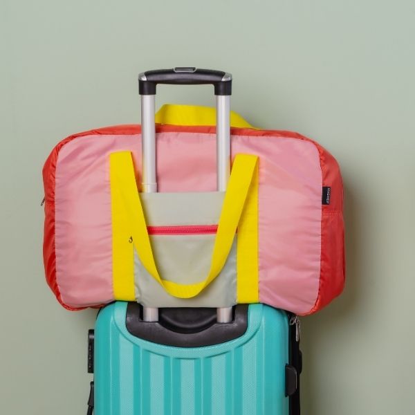 Un sac de voyage coloré, rose et rouge, muni de poignées jaunes, solidement fixé à la poignée télescopique d'une valise à coque rigide turquoise. La valise est posée devant un mur vert clair, prête à partir.