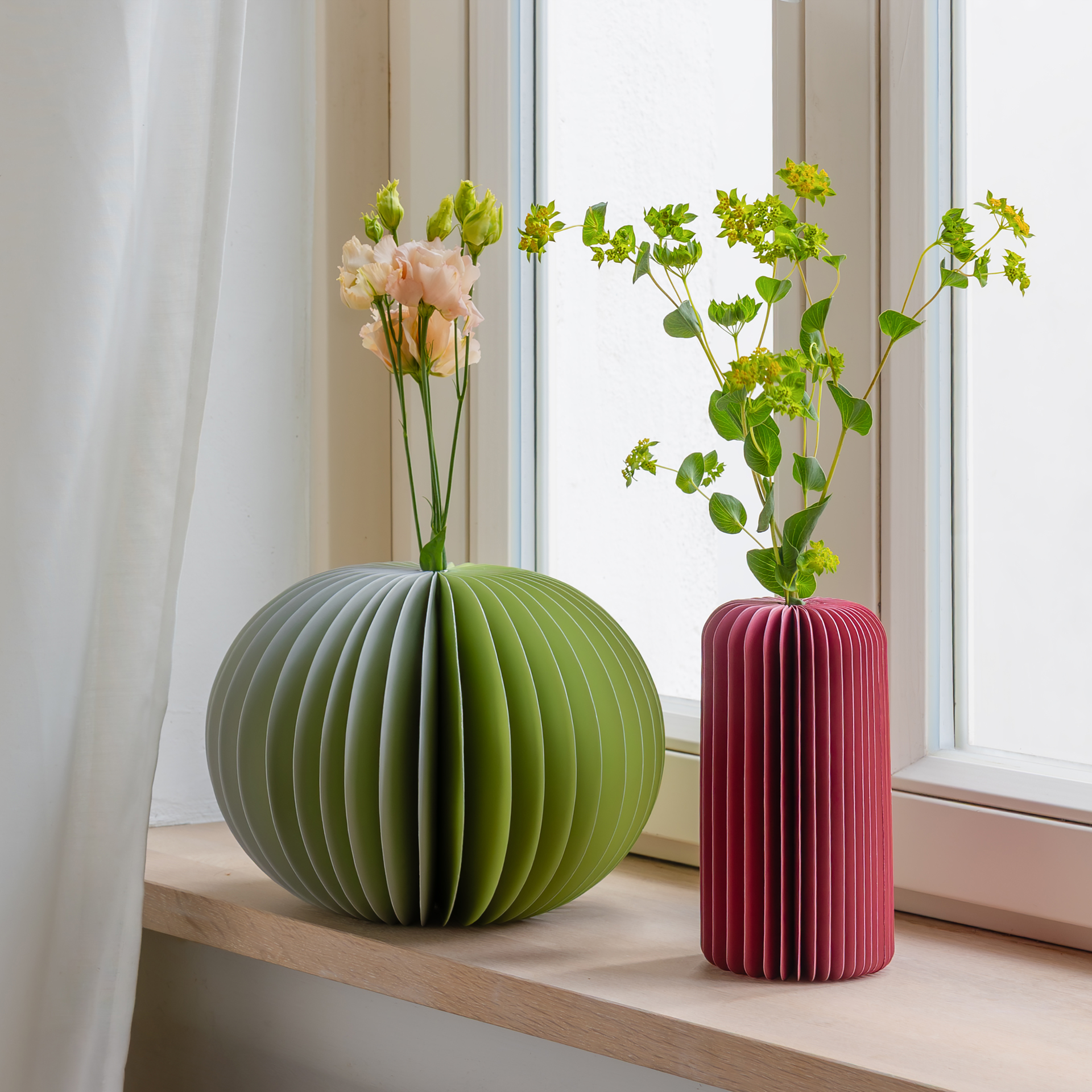 Zwei Papiervasen stehen auf einem Fensterbrett. Die linke Vase ist grün und rund, die rechte ist weinrot und zylindrisch. Beide enthalten Blumen: Pfirsichfarbene Rosen und grüne Euphorbia.