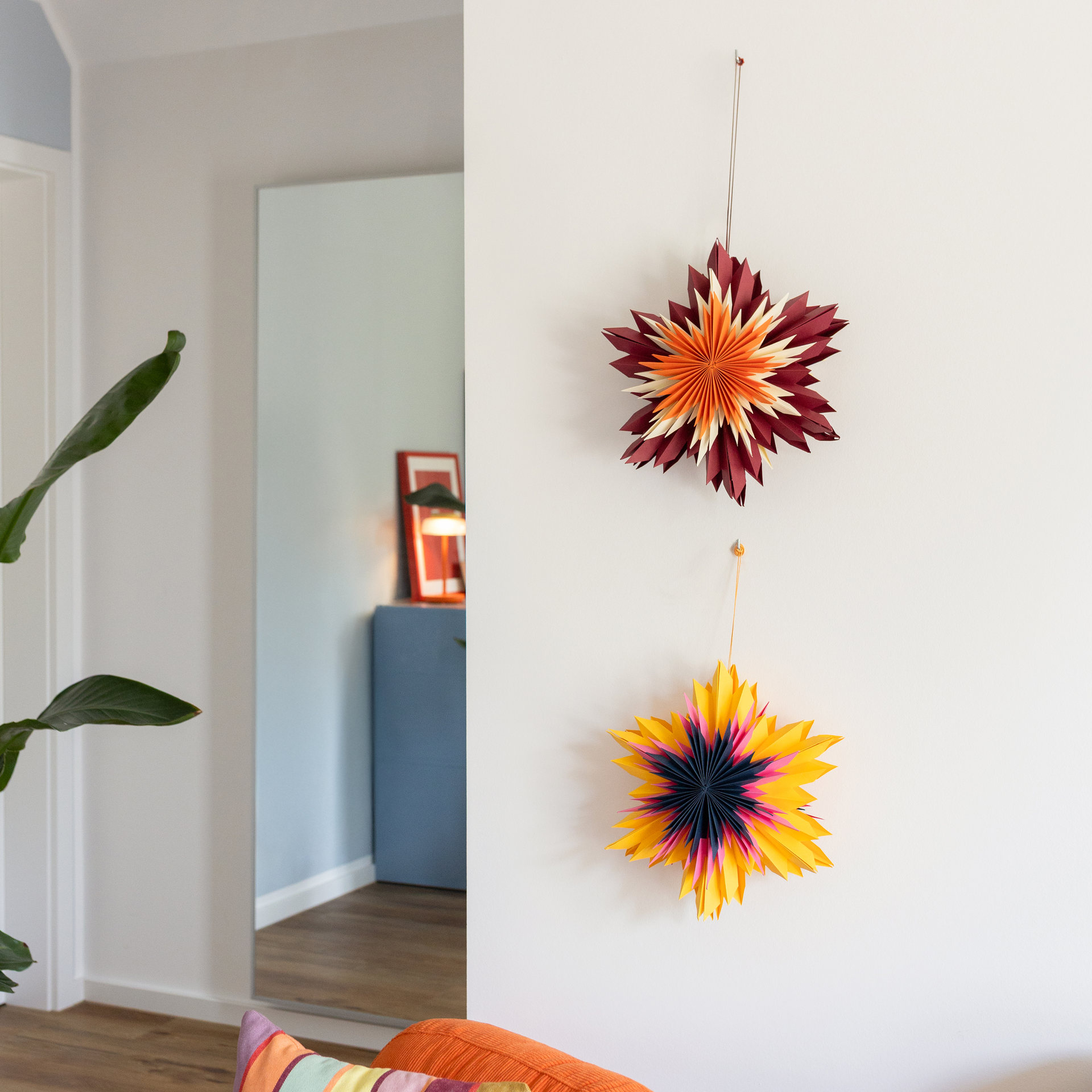 Deux étoiles décoratives en papier du lot Nova, avec boucle de suspension. À gauche, une étoile rouge foncé au centre orange-crème ; à droite, une étoile jaune vif au cœur bleu foncé-magenta. Motif plissé détaillé.