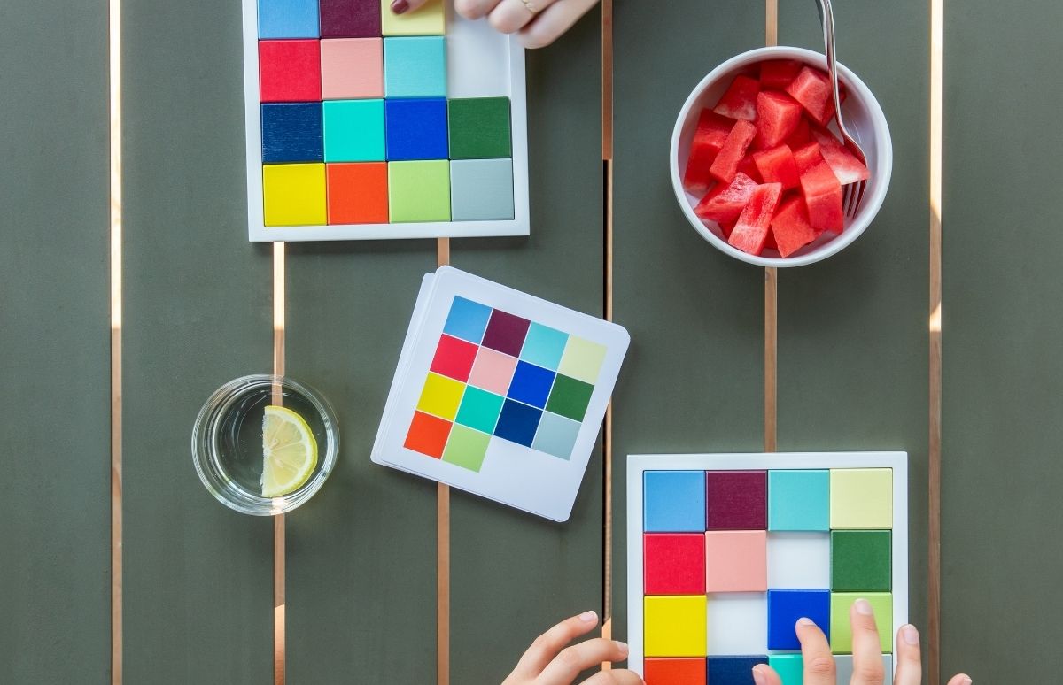 Deux mains disposent des blocs carrés colorés dans deux plateaux blancs. À côté, des piles de cartes, un verre d'eau au citron et une assiette de pastèque sur une table verte.