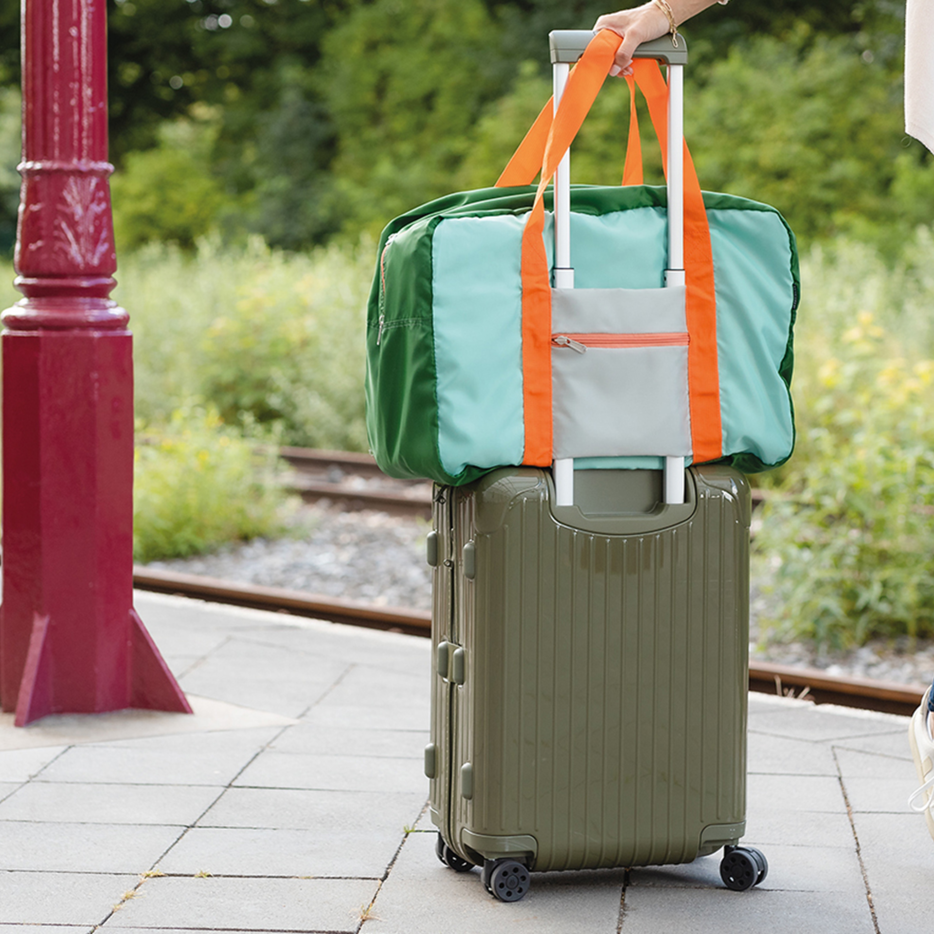 Eine grüne Reisetasche 'Max' mit orangefarbenen Trägern liegt auf einem olivgrünen Rollkoffer. Beide stehen auf einem Bahnsteig, im Hintergrund ist Grün zu sehen.