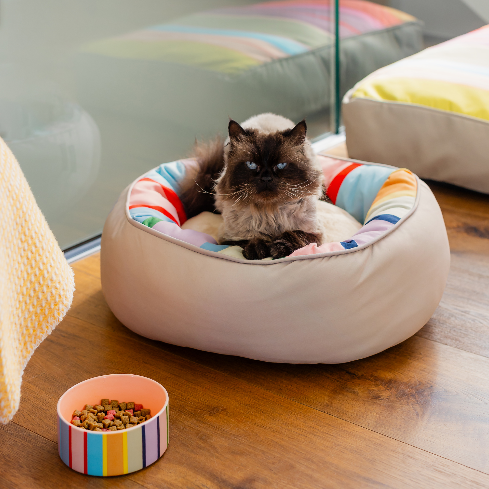 Lit rond pour chiens et chats Lotta S, taille S, avec un revêtement extérieur lisse de couleur beige. À l’intérieur, un coussin moelleux et amovible orné de rayures colorées en jaune, orange, bleu, rose et vert pour un confort optimal.