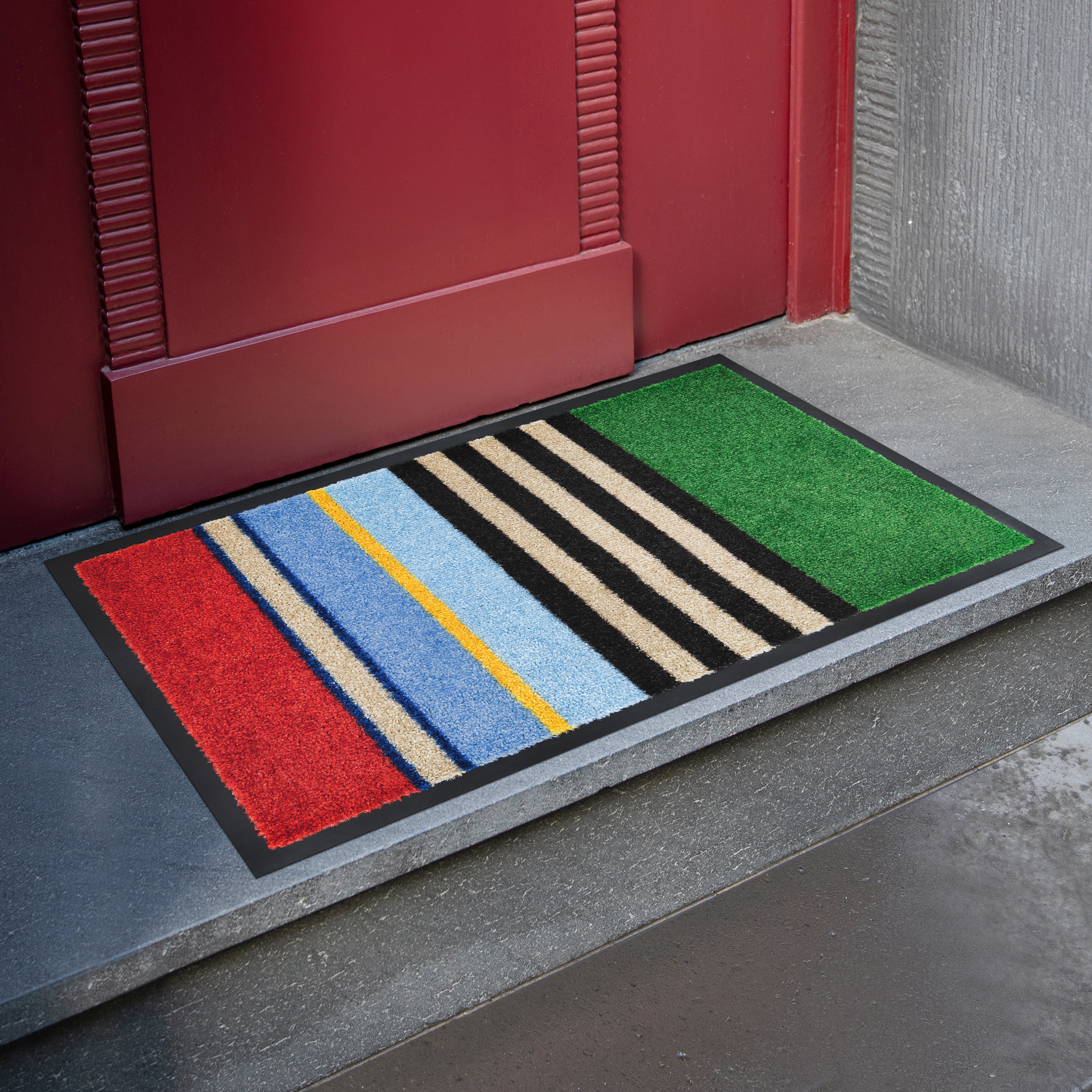 Ein rechteckiges, buntes Bodenaccessoire liegt vor einer roten Tür auf einem dunklen Steinboden. Das Bodenaccessoire weist schräge Streifen in rot, beige, blau, gelb, schwarz und grün auf.