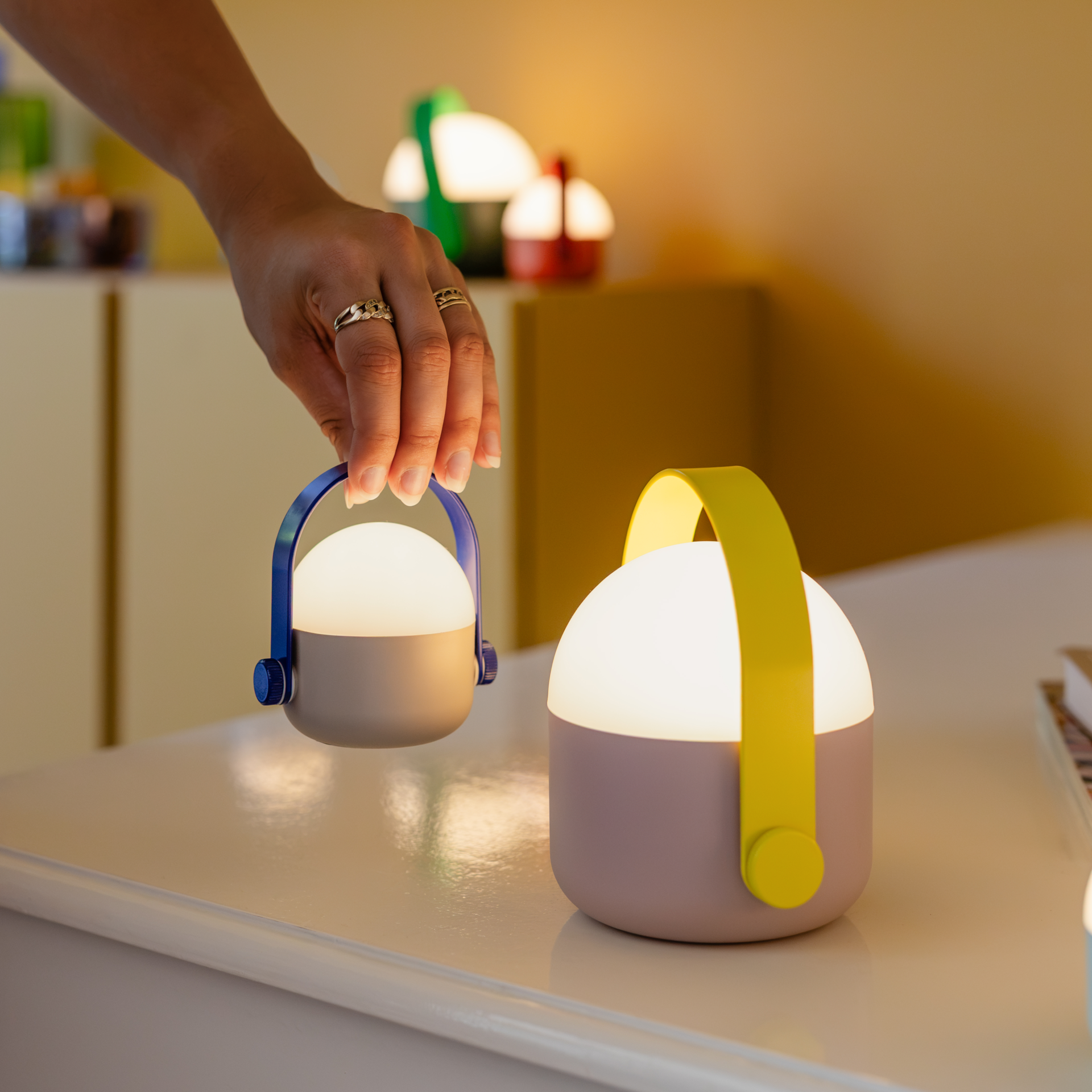 Una mano regge una lampada LED OLE mini di colore blu-grigio, accesa. Accanto ad essa, su una superficie chiara, si trova una lampada OLE mini giallo-rosa, anch’essa accesa. Sullo sfondo si intravedono sfocate altre due lampade. Luce bianca calda.