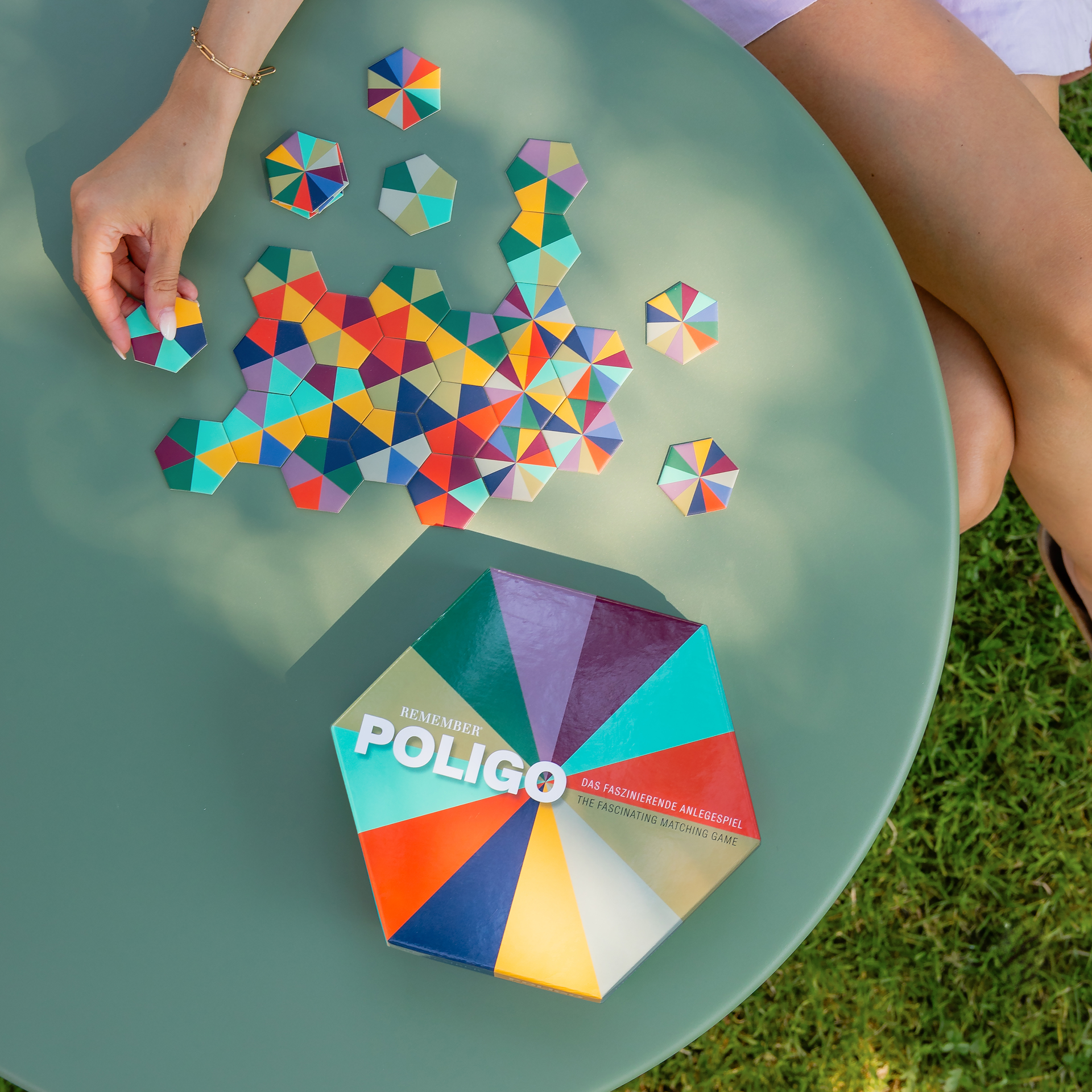Draufsicht auf Poligo Anlegespiel: Bunte, sechseckige Spielkarten liegen auf einem grünen Tisch, teilweise zu einem Muster gelegt. Eine Hand hält eine Karte. Die Poligo Spielbox ist sichtbar.