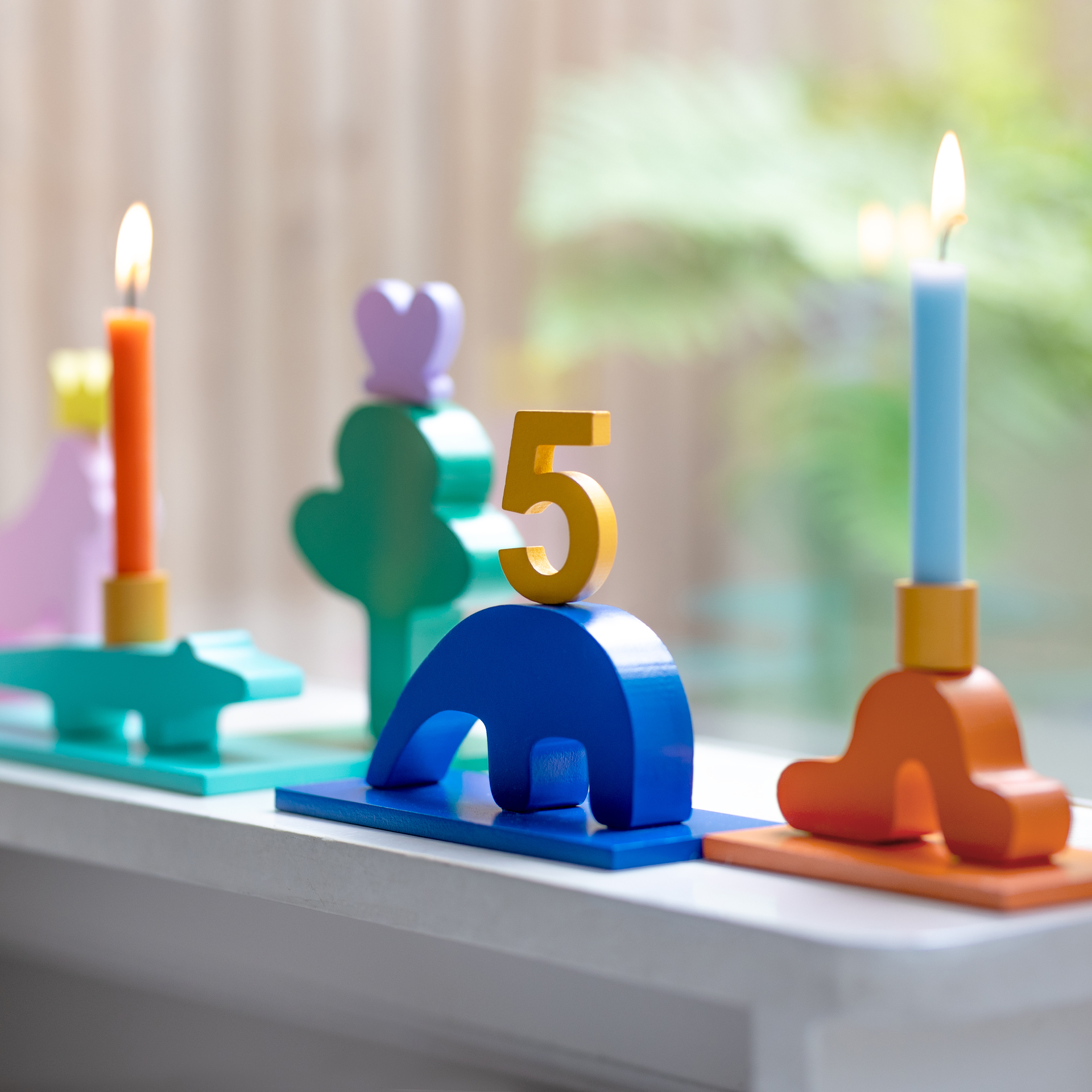 Bunte, hölzerne Geburtstagsdekoration mit zwei brennenden Kerzen. In der Mitte steht eine goldene „5“ auf einem blauen Bogen. Formen in Grün, Lila, Rosa und Orange akzentuieren.