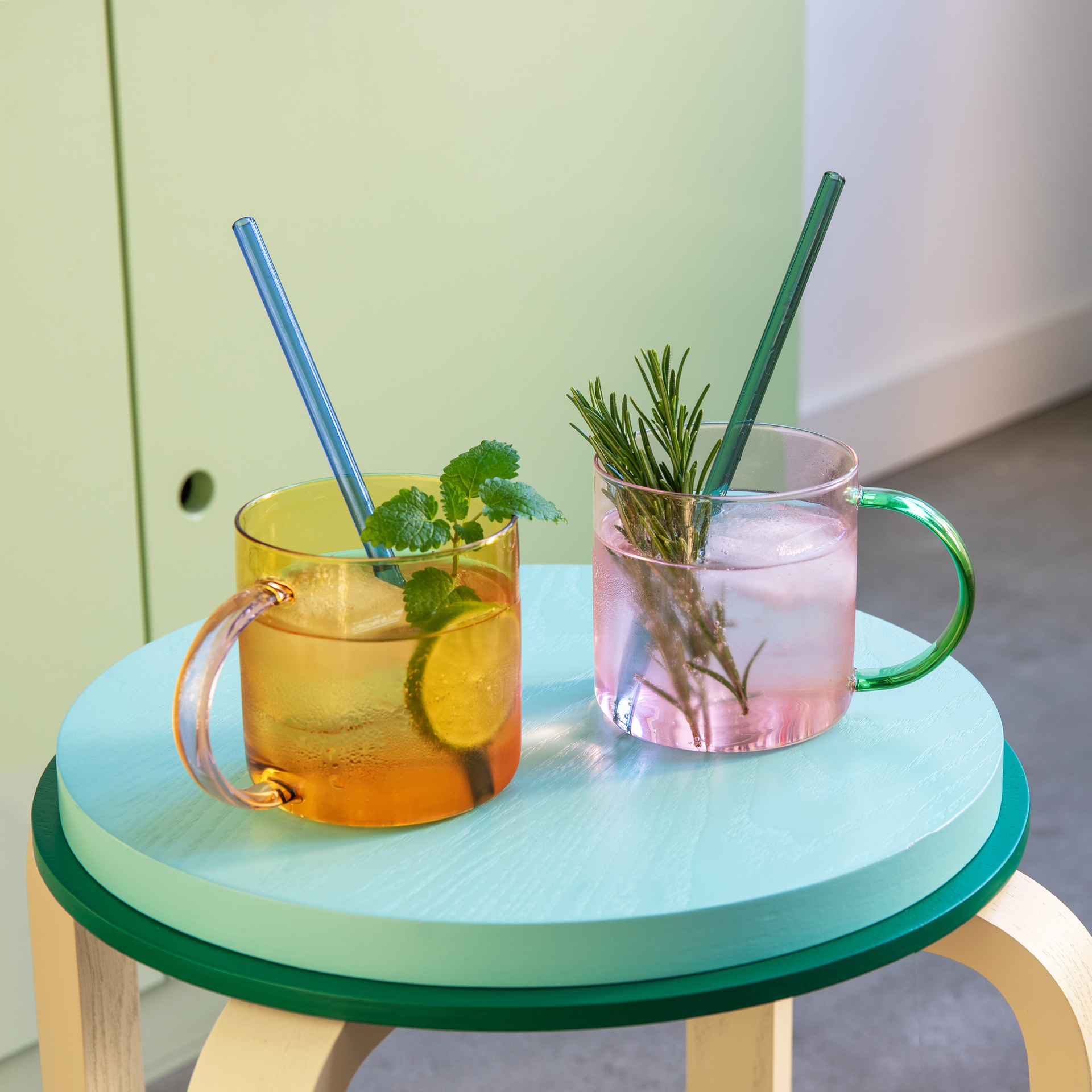Zwei Glasbecher 'Fred', einer rosefarben mit grünem Henkel, der andere gelb mit rosafarbenem Henkel, stehen nebeneinander vor einem weißen Hintergrund.