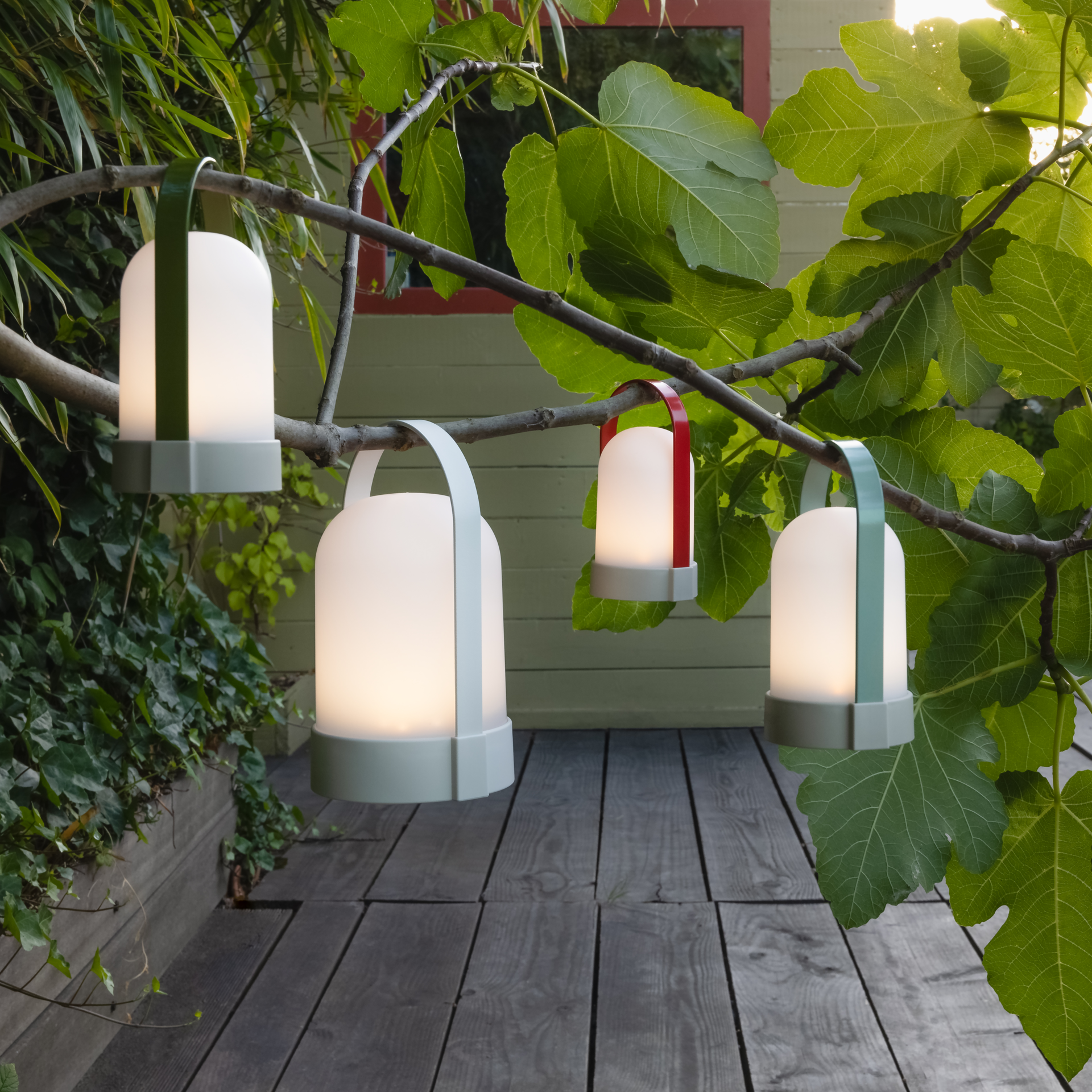 Im Garten hängen vier URI Leuchten an einem Ast, der von links nach rechts verläuft. Die Leuchten spenden warmes Licht und die Tragebügel sind farblich unterschiedlich.