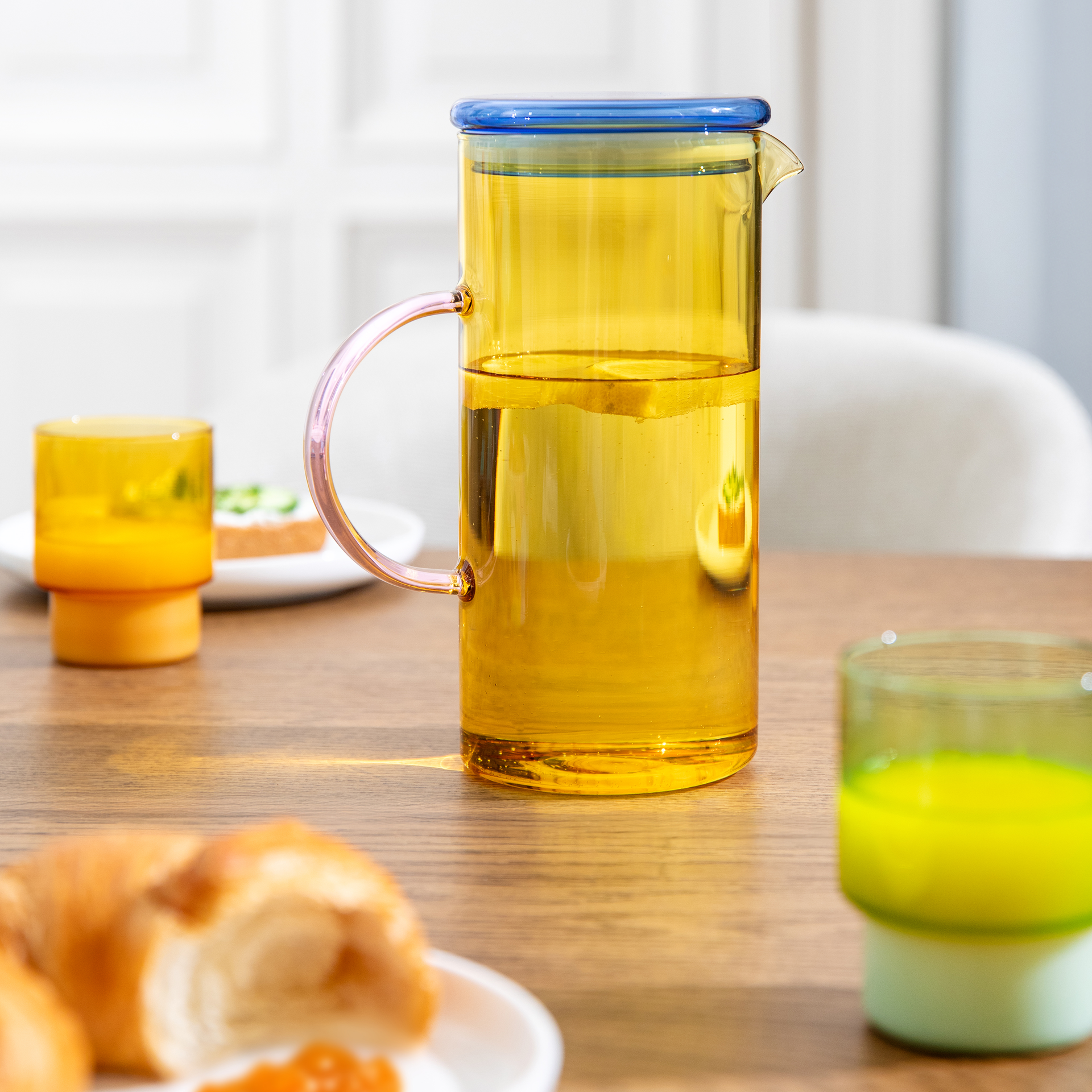 Un tavolo con colazione. Il centrotavola è una caraffa di vetro giallo con coperchio blu e manico rosa, riempita di liquido e fette di limone. Il tutto viene servito con un croissant, marmellata e bevande.
