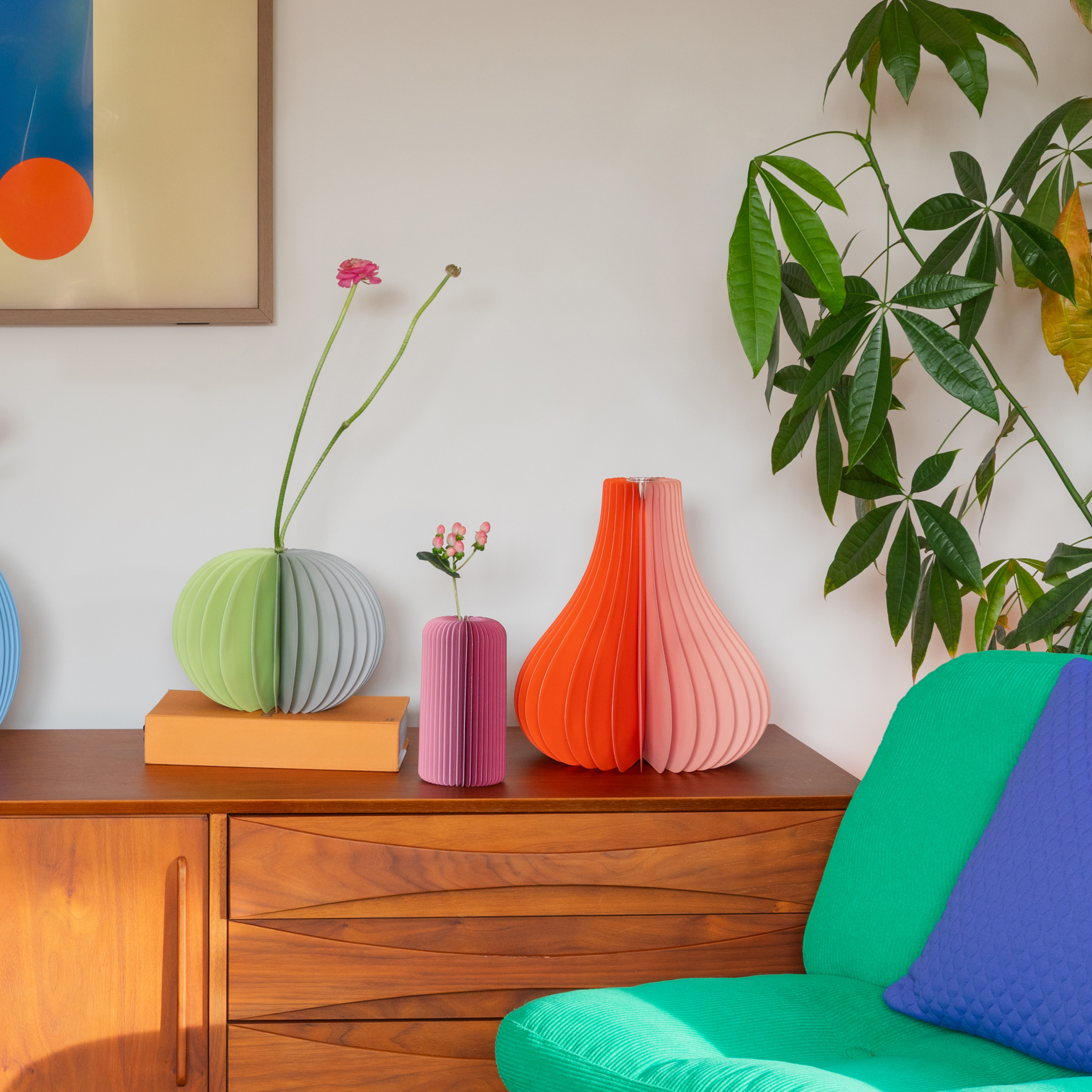 Auf einem Holzschrank stehen bunte Vasen mit Blumen. Daneben eine grüne Pflanze und ein Sessel mit grünem Kissen. Im Hintergrund ein abstraktes Bild.