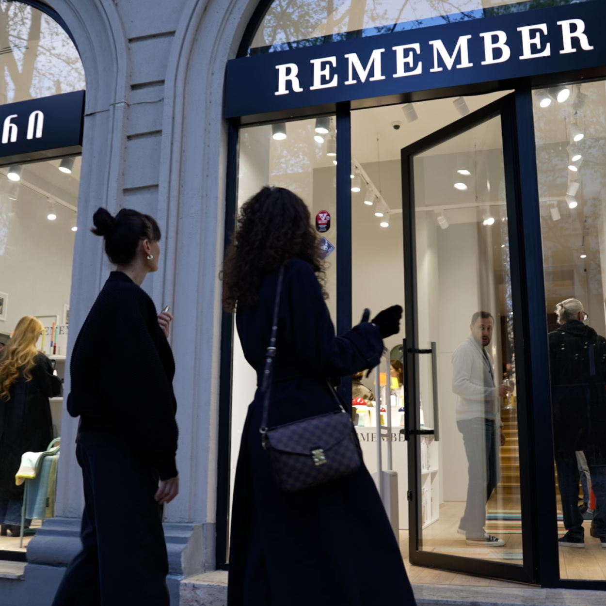 Zwei Frauen vor einem Geschäft mit der Aufschrift „Remember“. Eine Frau trägt einen dunklen Mantel mit Handtasche, die andere eine dunkle Jacke. Personen sind durch die offene Tür des Geschäfts zu sehen.