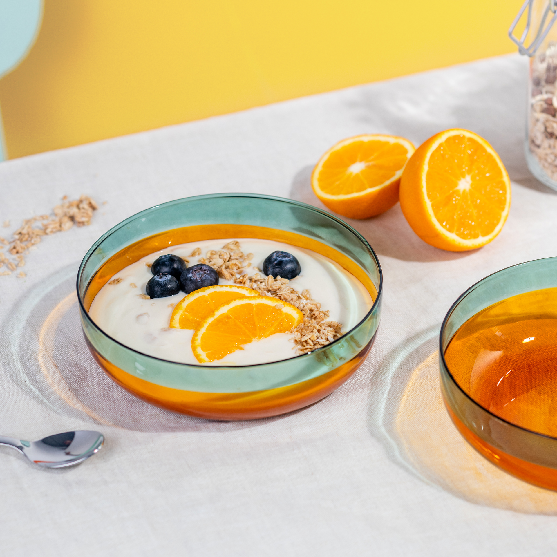 Zwei Glasschälchen 'amber' auf Stoff. Eins gefüllt mit Joghurt, Blaubeeren, Orange, Müsli; daneben halbe Orangen.