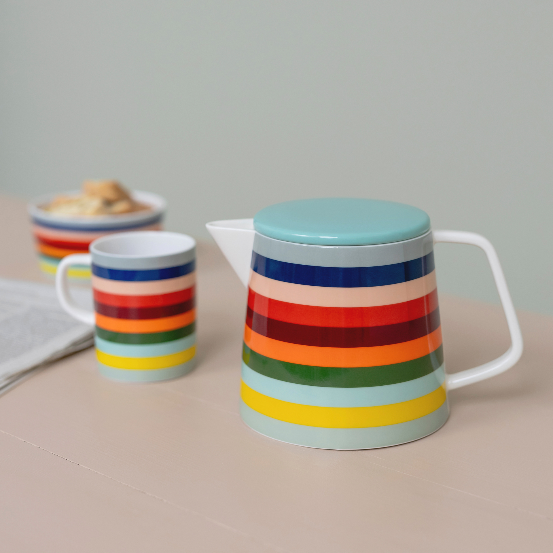 Eine 'Lorenzo' Teekanne mit türkisem Deckel und bunten Streifen, daneben eine Tasse im gleichen Design und eine Schale mit Gebäck.