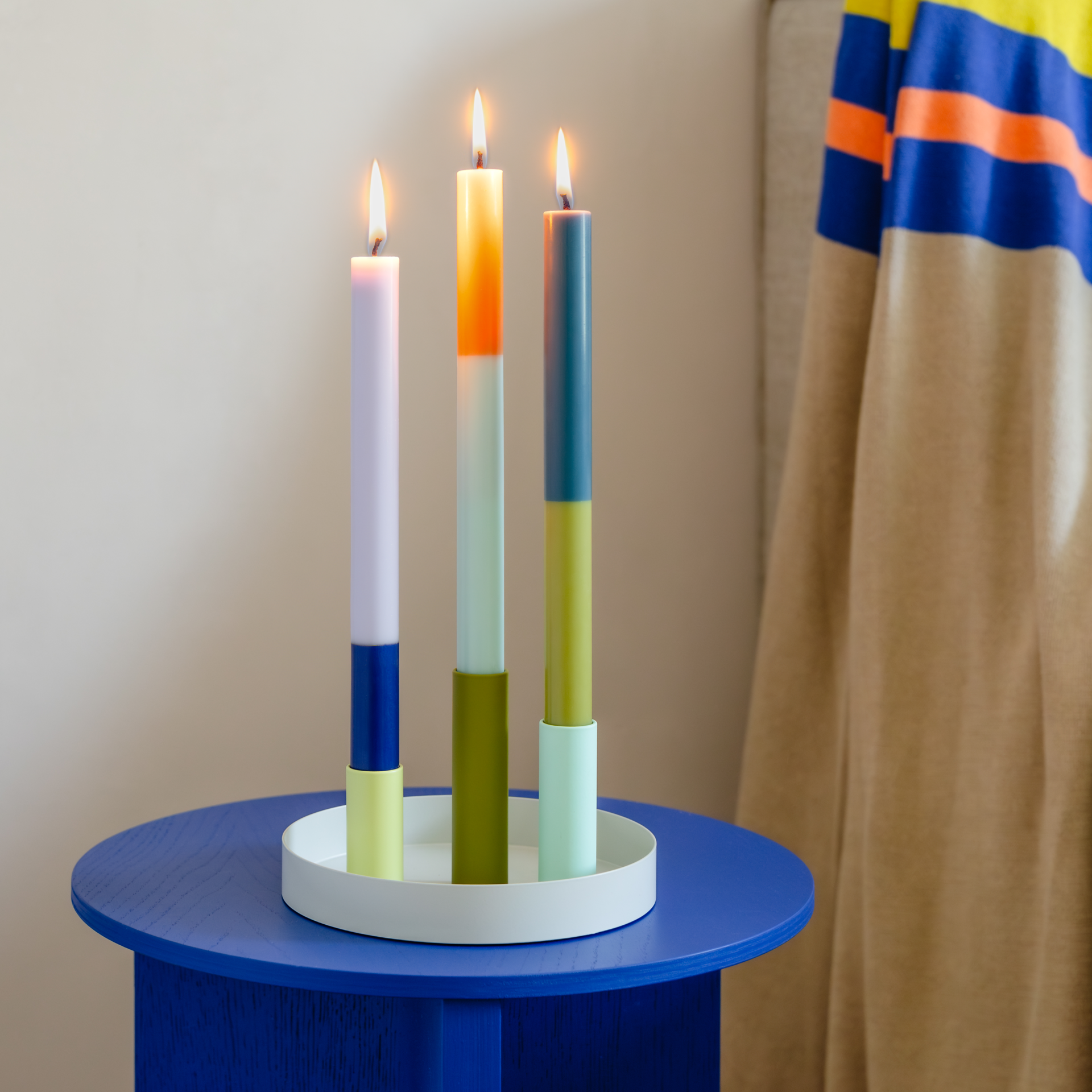 Drei bunte Stabkerzen im 'Luna Mini' Kerzenhalter-Set stehen auf einem blauen Tisch. Die Kerzenhalter sind farblich passend zu den unteren Hälften der Kerzen. Alle Kerzen sind angezündet.
