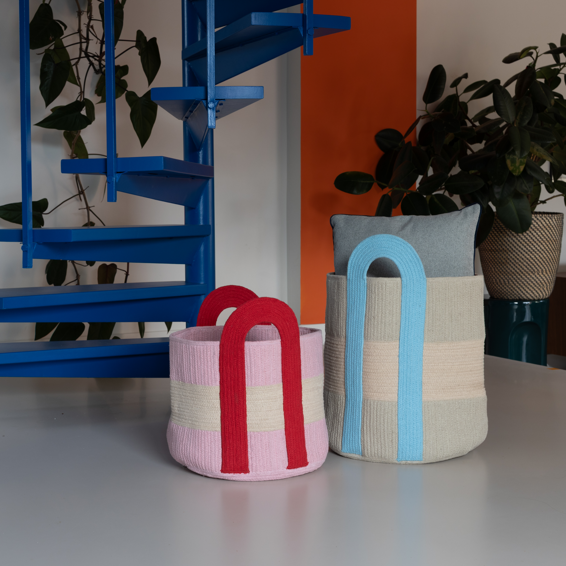 Zwei große, zylinderförmige 'Colombo' Körbe aus Baumwolle, der eine rosa mit roten Henkeln, der andere grau mit blauen Henkeln, im Interieur.