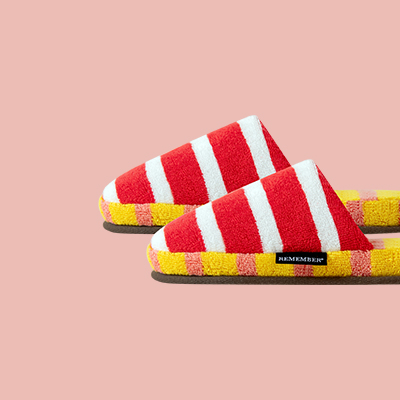 Zwei flauschige, offene Hausschuhe mit roten und weißen Streifen, gelbem Rand und dunkelbrauner Sohle, auf einem pinkfarbenen Hintergrund. Ein kleines schwarzes Etikett mit weißer Schrift ist zu sehen.
