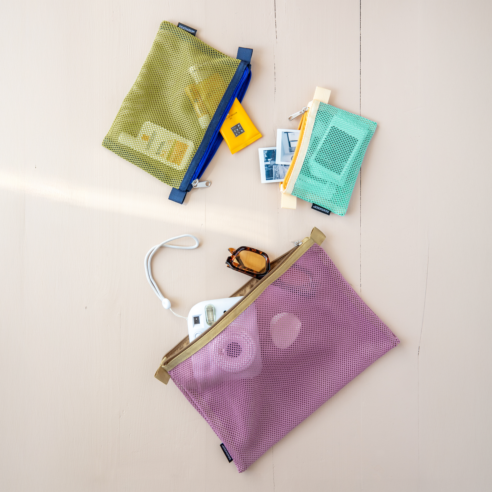Drei farbige Mesh Bag Sets, offen auf einer Fläche. Die Farben sind Grün, Türkis und Lila. In den Taschen sind Kosmetik, Sonnenbrille und eine Kamera.