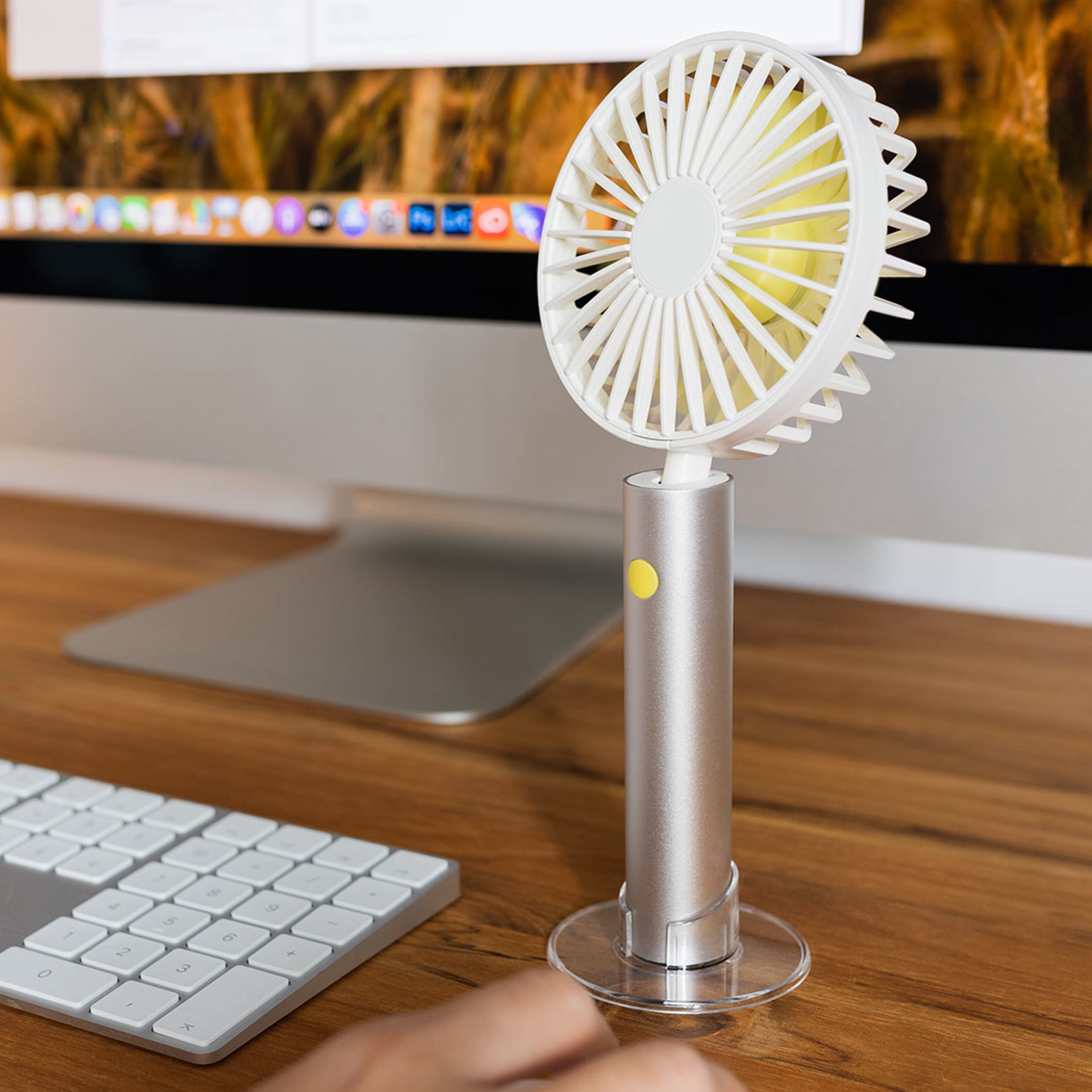 Ein silberner, aufladbarer Ventilator steht auf einem Schreibtisch neben einer Tastatur. Der Ventilator hat einen transparenten Standfuß und einen weißen Rotorkopf. Im Hintergrund ist ein Computerbildschirm zu sehen.