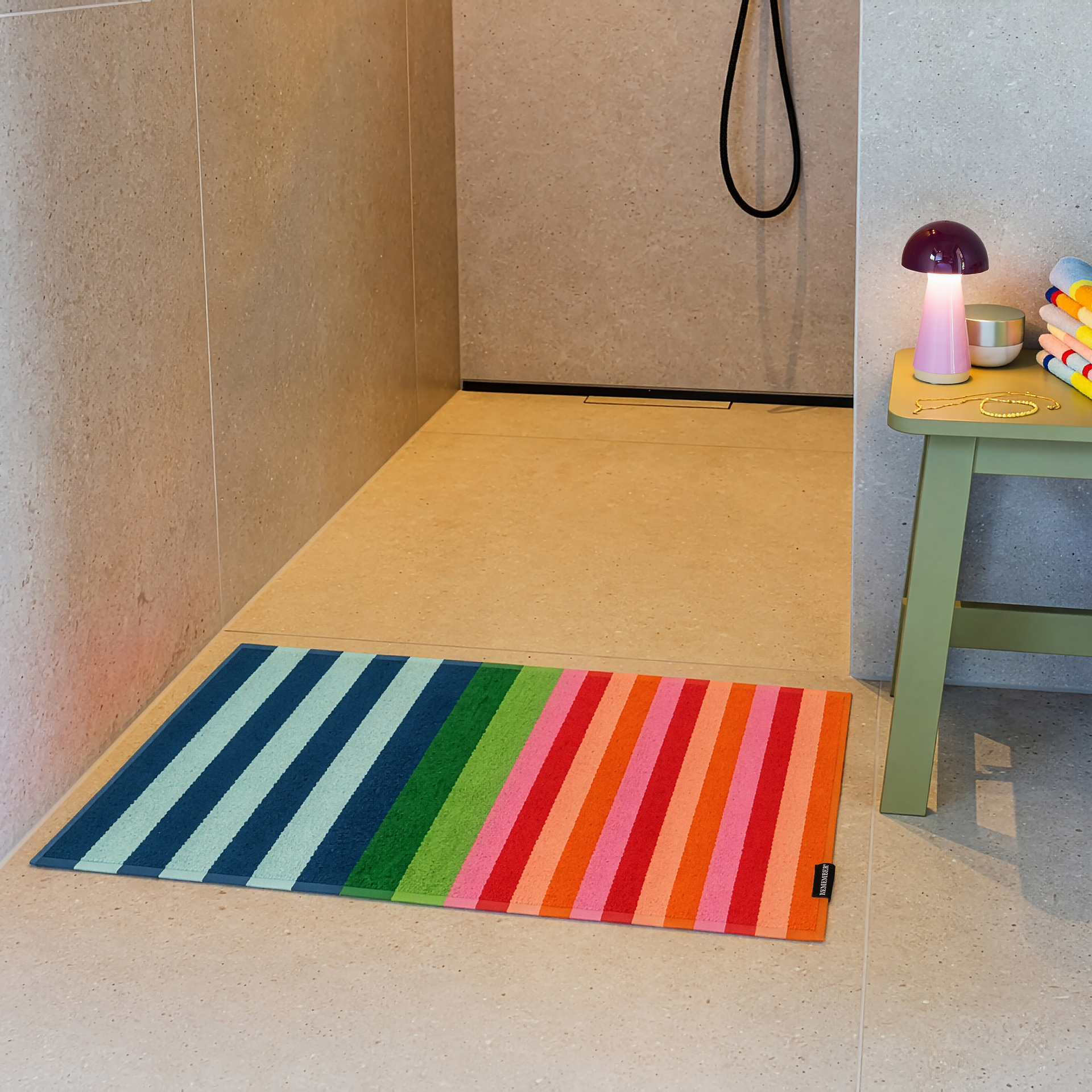 Tapis de bain coloré « Milo » à rayures verticales multicolores : à gauche, alternance de bleu clair et de bleu foncé ; au centre, vert et vert clair ; à droite, rayures vives en rouge, rose et orange. Petite étiquette noire en bas à droite.