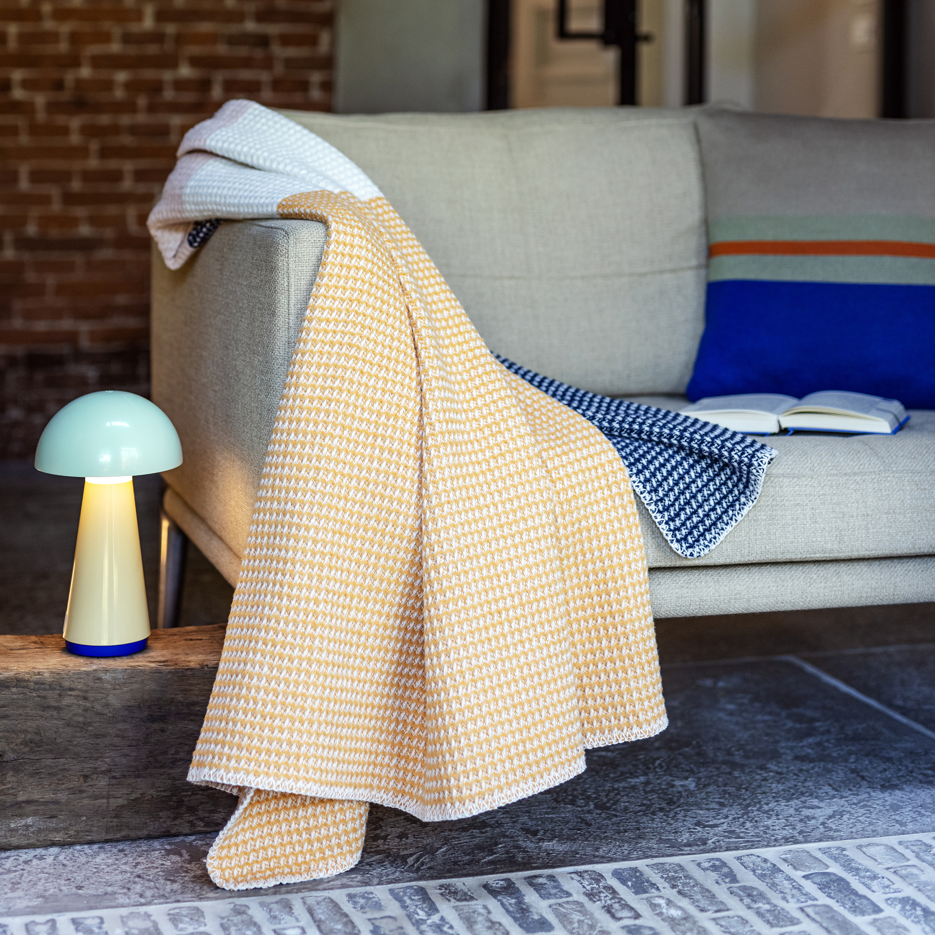 Beige Couch mit Zierkissen und aufgeschlagenes Buch. Die Gala-Decke in Gelb hängt über die Couchlehne. Daneben steht eine gelbe Lampe.