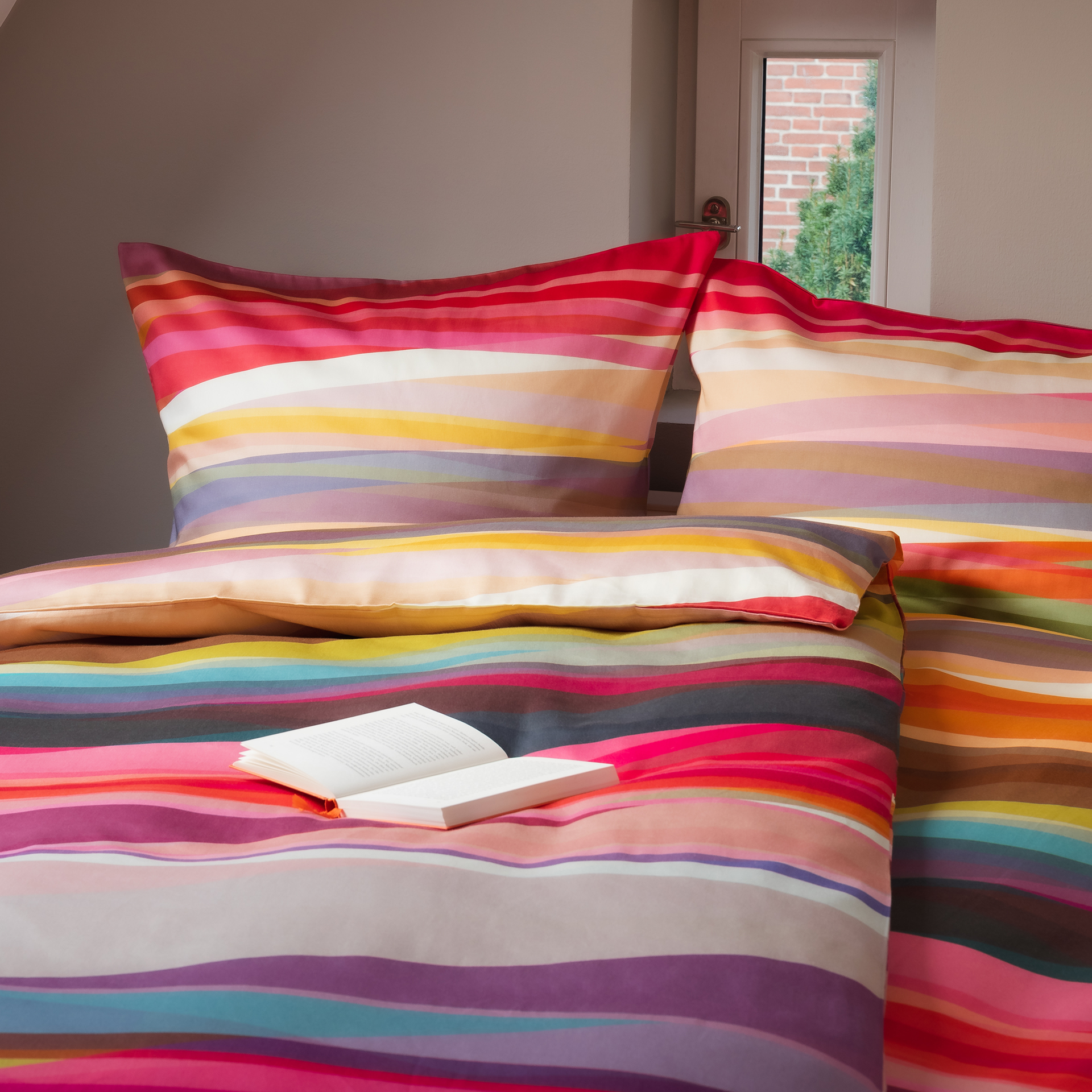 Detailaufnahme: gestreifte Bettwäsche 'Mateo' aus Mako-Satin mit Buch auf der Decke, zwei Kissen und Fenster im Hintergrund.