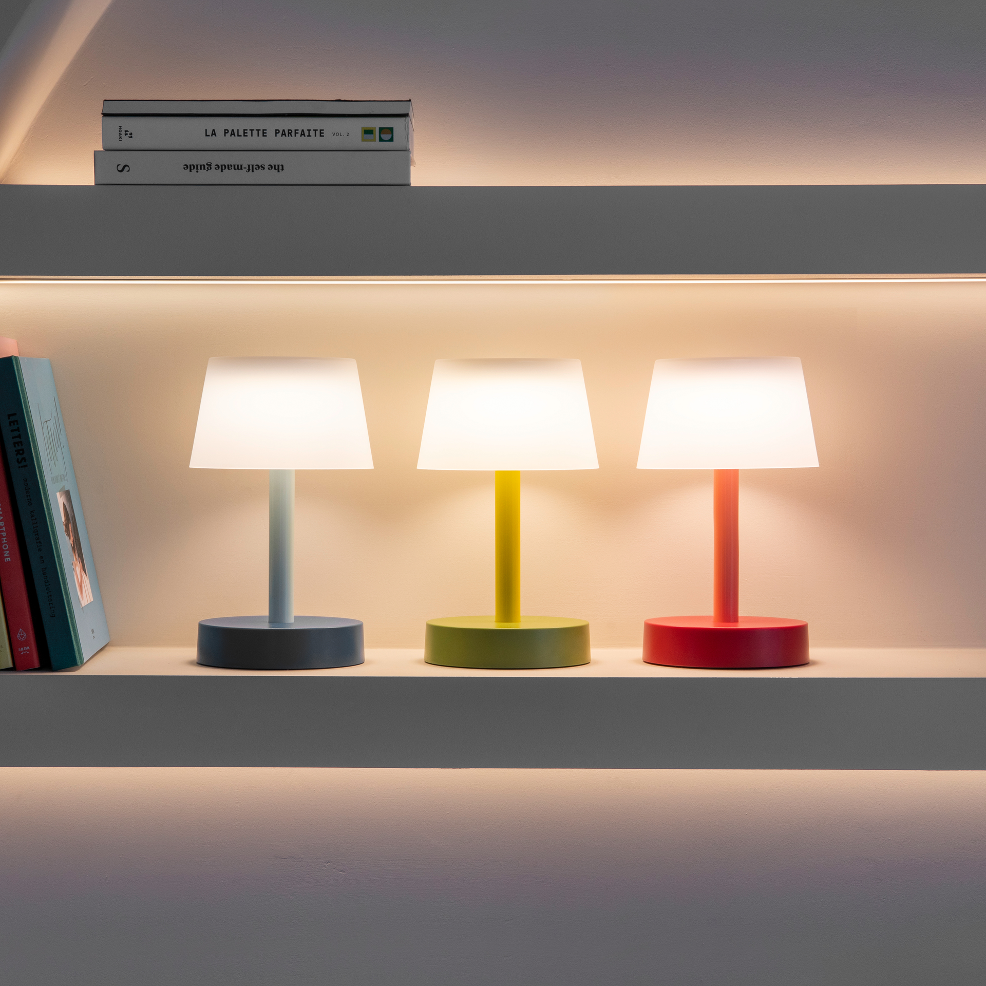 Drei farbige Fritz Tischleuchten im Regal-Setup. Die Leuchten in blau, gelb und rot sind aktiviert und emittieren warmes Licht. Bücher sind nebenan und darüber platziert.